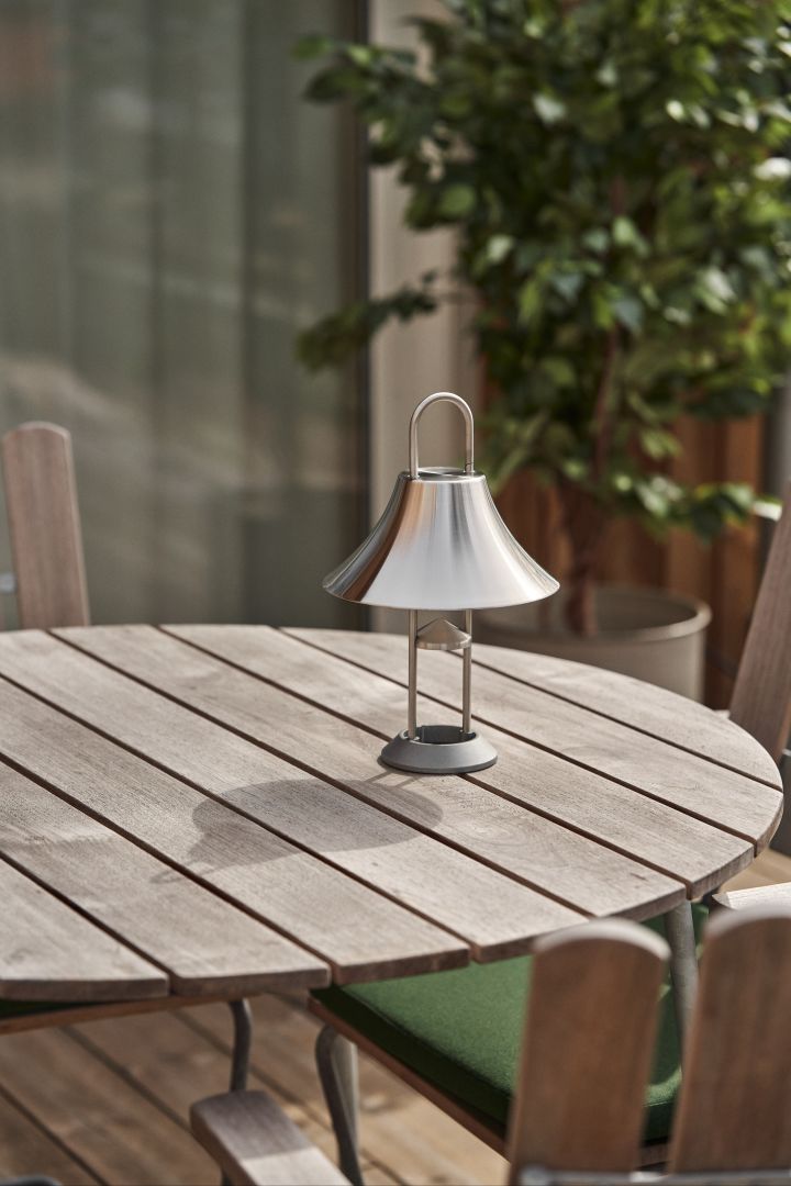 Bring atmospheric light to your garden or patio with the Mousqueton portable lamp in stainless steel from HAY, standing on a round teak outdoor table.