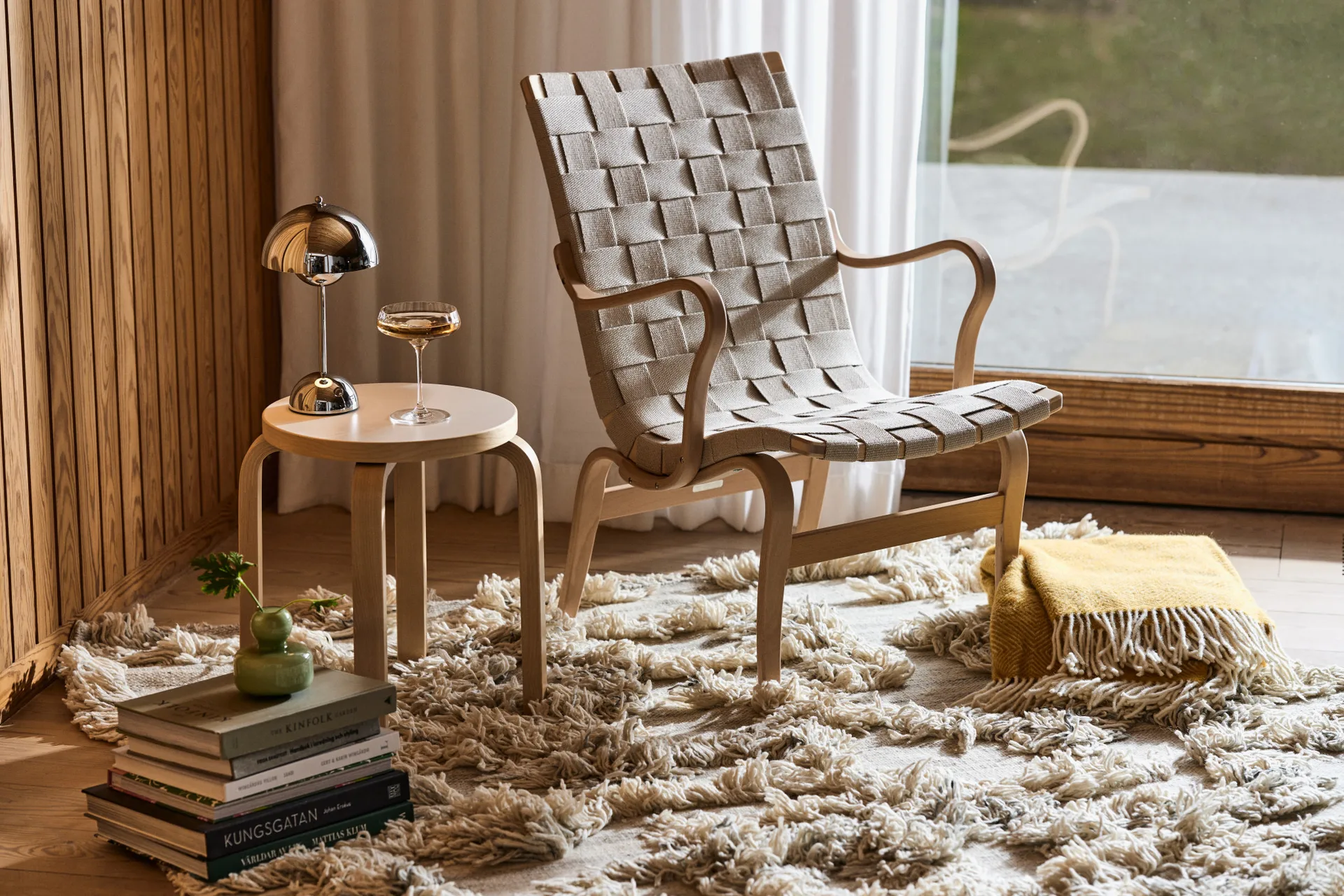 Light wood lounge chair with woven beige fabric on a shaggy rug, next to a table with a chrome lamp and a drink. Books nearby.