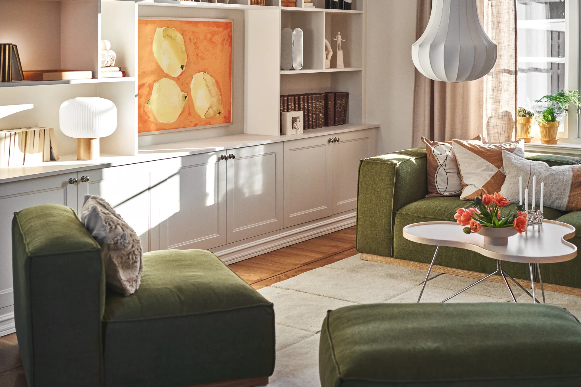 Sunlit living room with green sofas, a white coffee table with orange tulips and candles, and built-in white shelves with art.