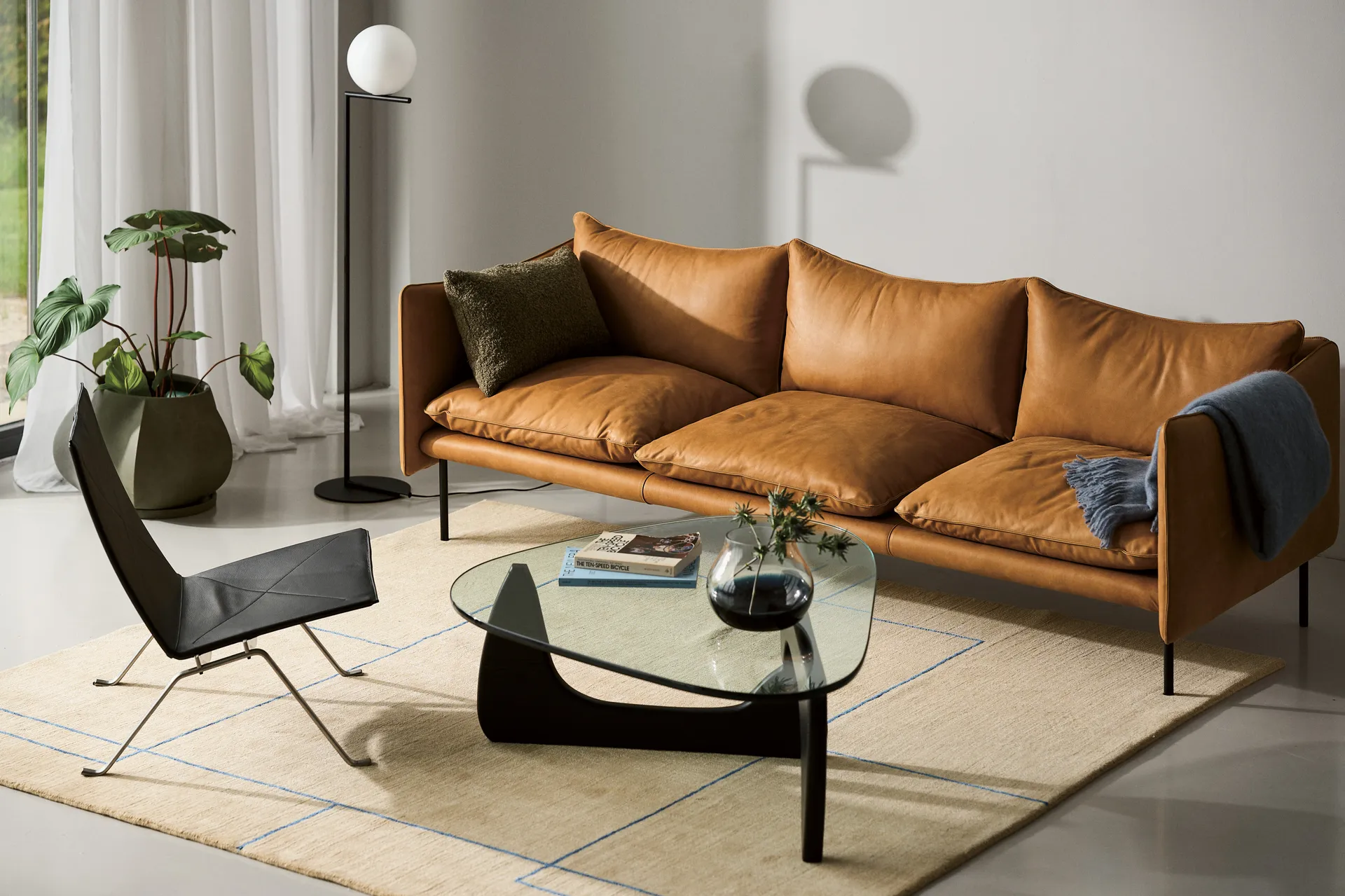 A modern living room with a tan leather sofa, black lounge chair, glass coffee table, and a beige rug with blue lines.