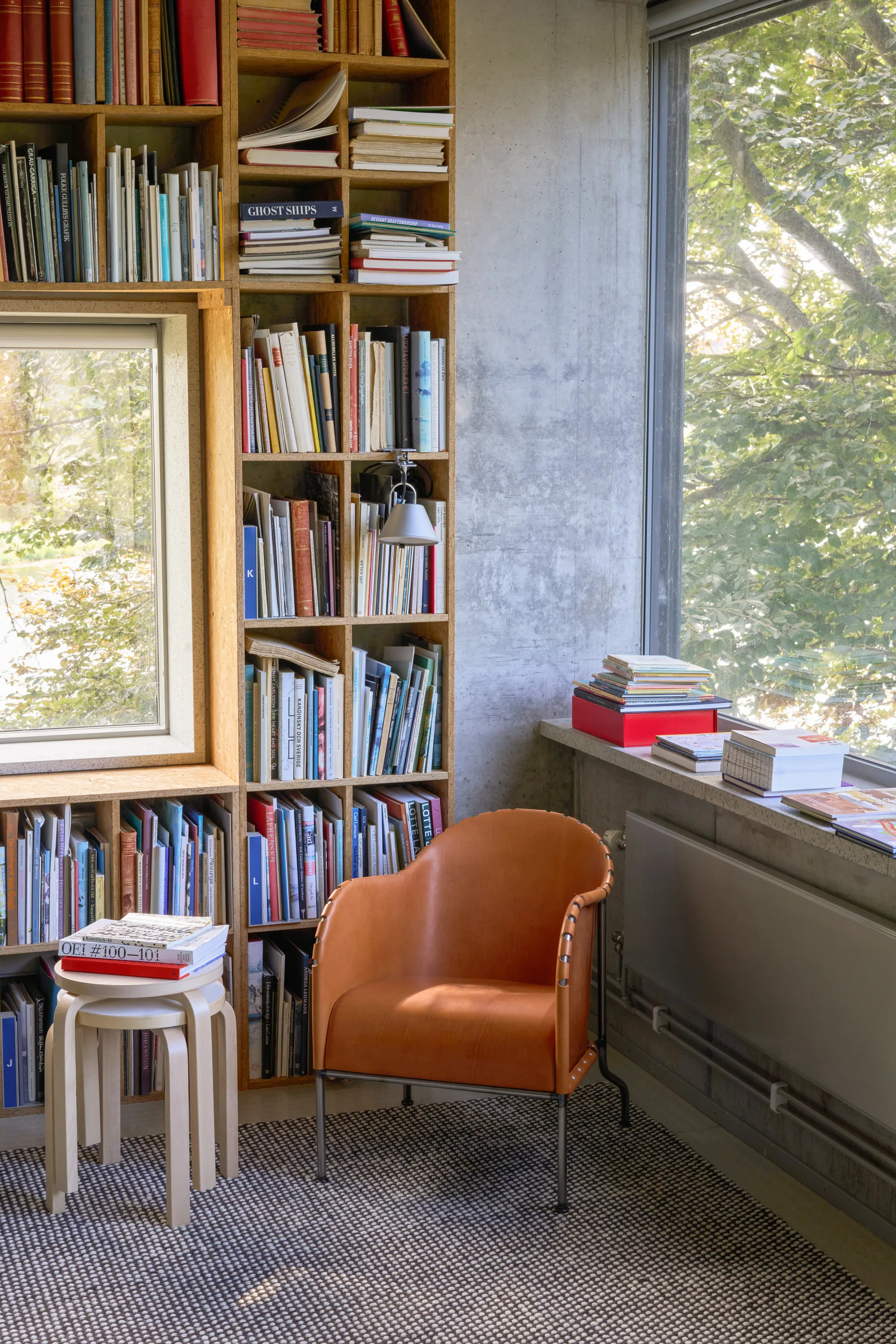 A cozy room with a large wooden bookshelf, a light brown leather armchair, a small stool with books, and a window looking out to trees.