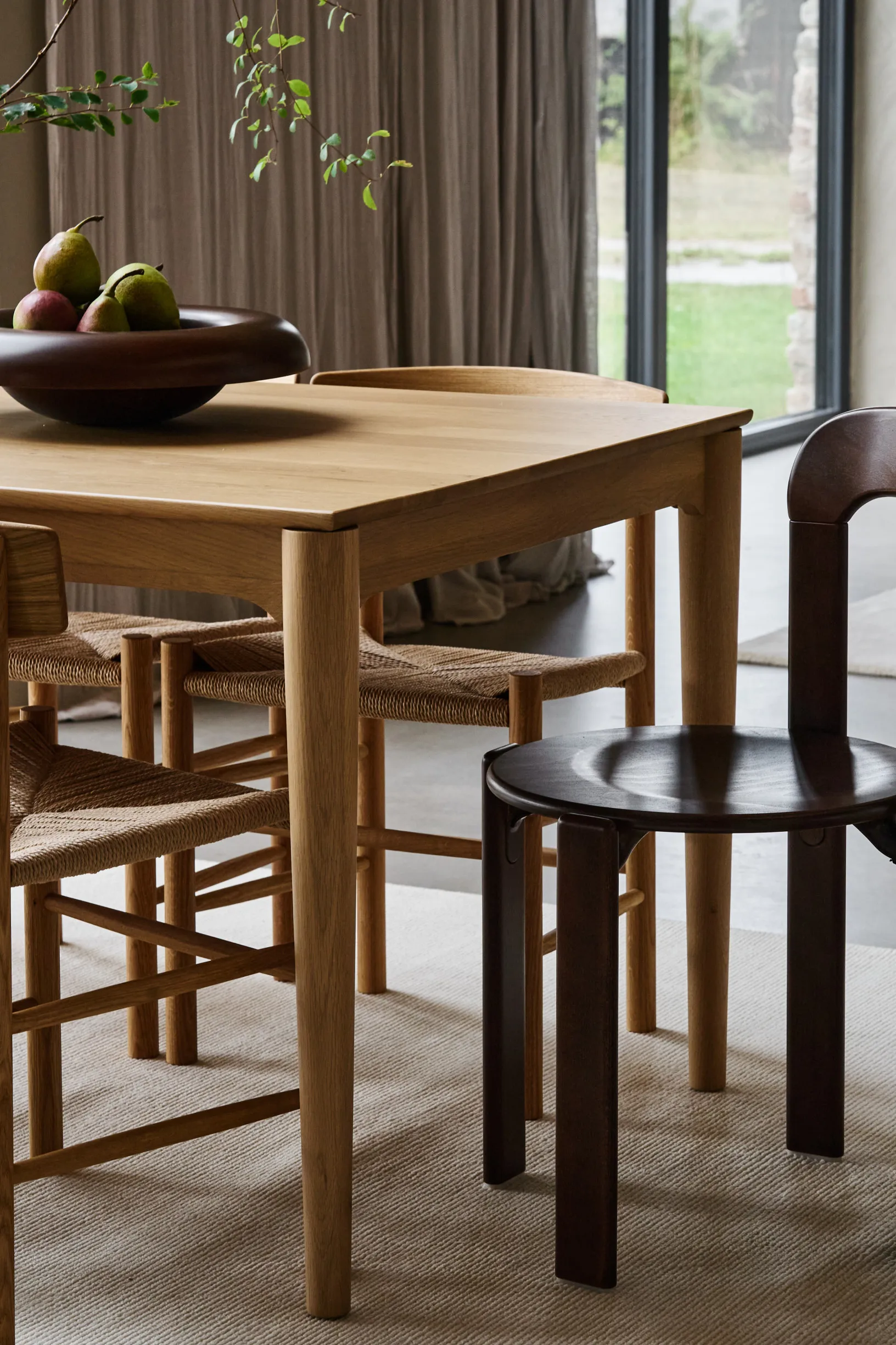 Wooden dining table with a fruit bowl, light wood chairs with woven seats, and a dark wood chair on a rug.