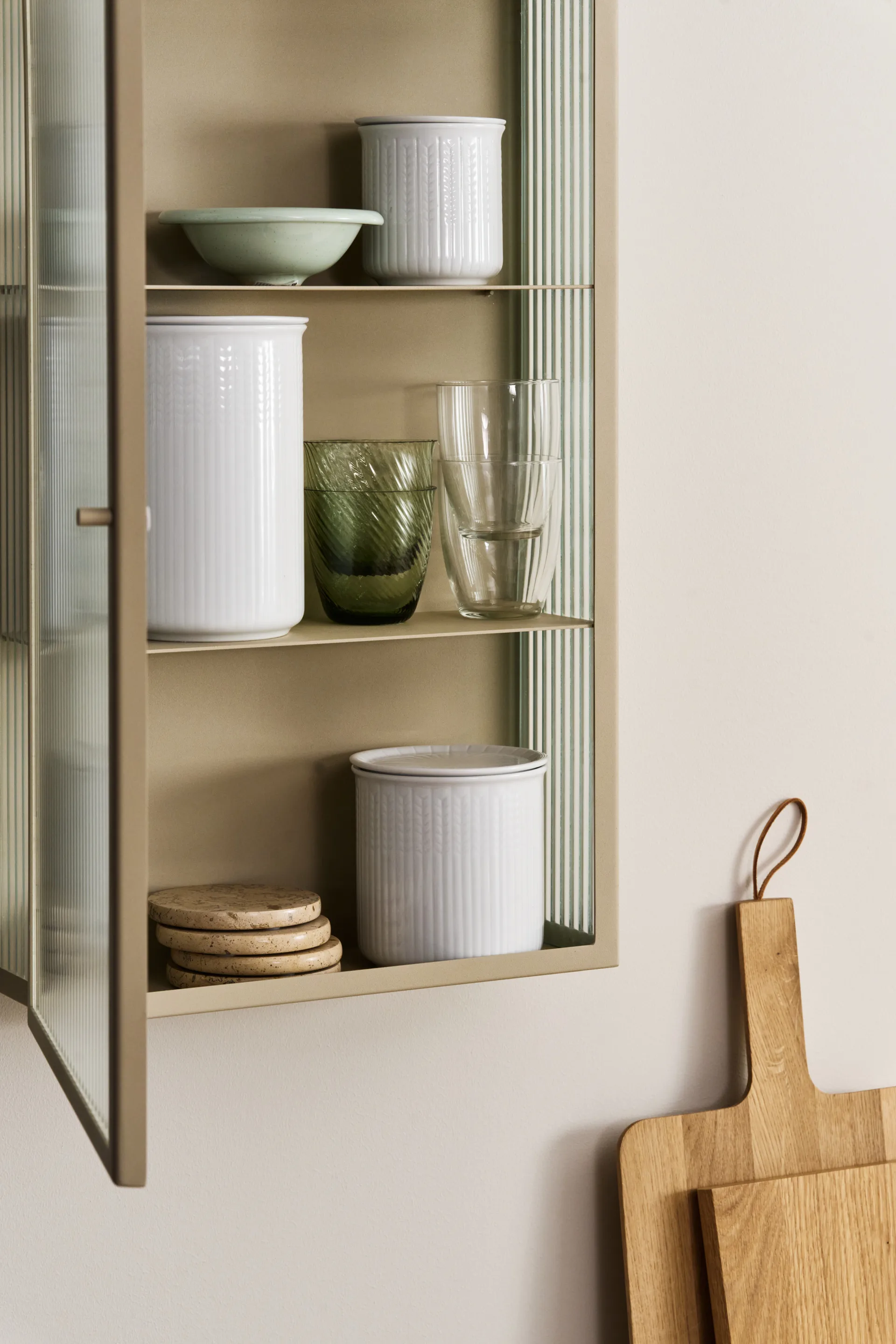 A light beige cabinet with ribbed glass doors, open to reveal white containers, green and clear glasses, and coasters. Wooden cutting boards hang nearby.