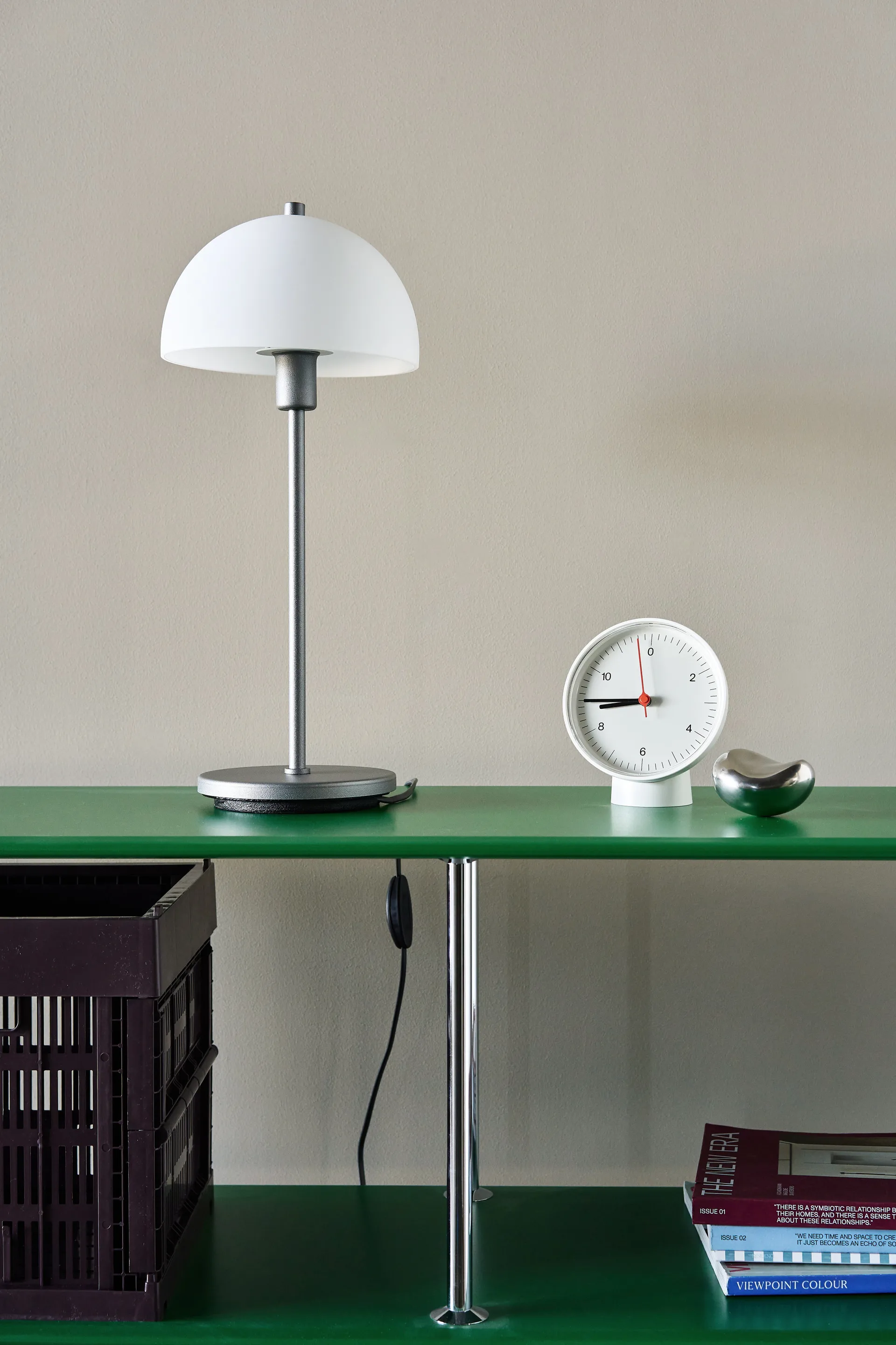 Minimalist green shelf with a white dome lamp, a white round clock, and a silver pebble. A dark crate and magazines are below.