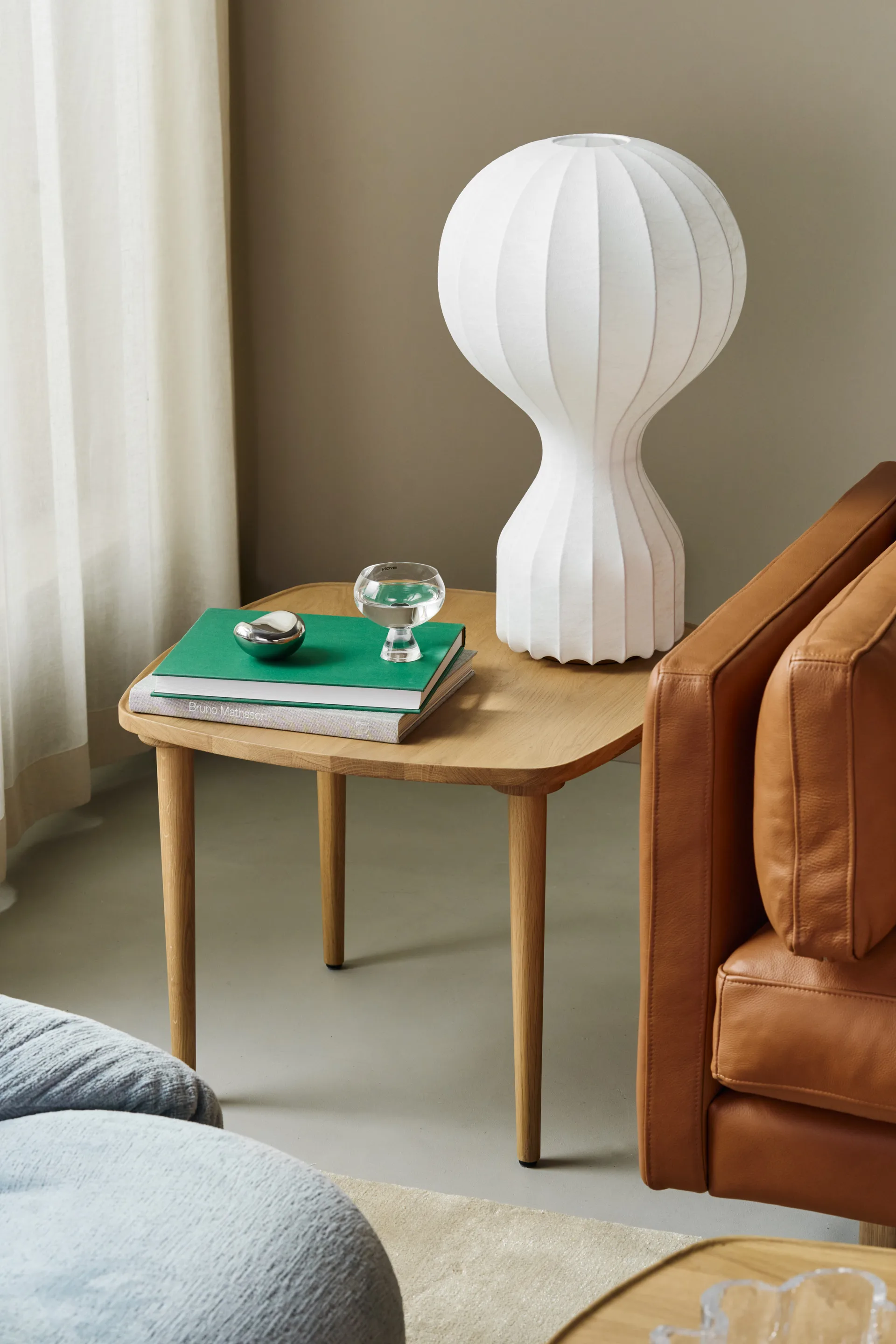 Light wood side table with a white ribbed lamp, green books, a glass of water, and a silver object, next to a brown leather chair.