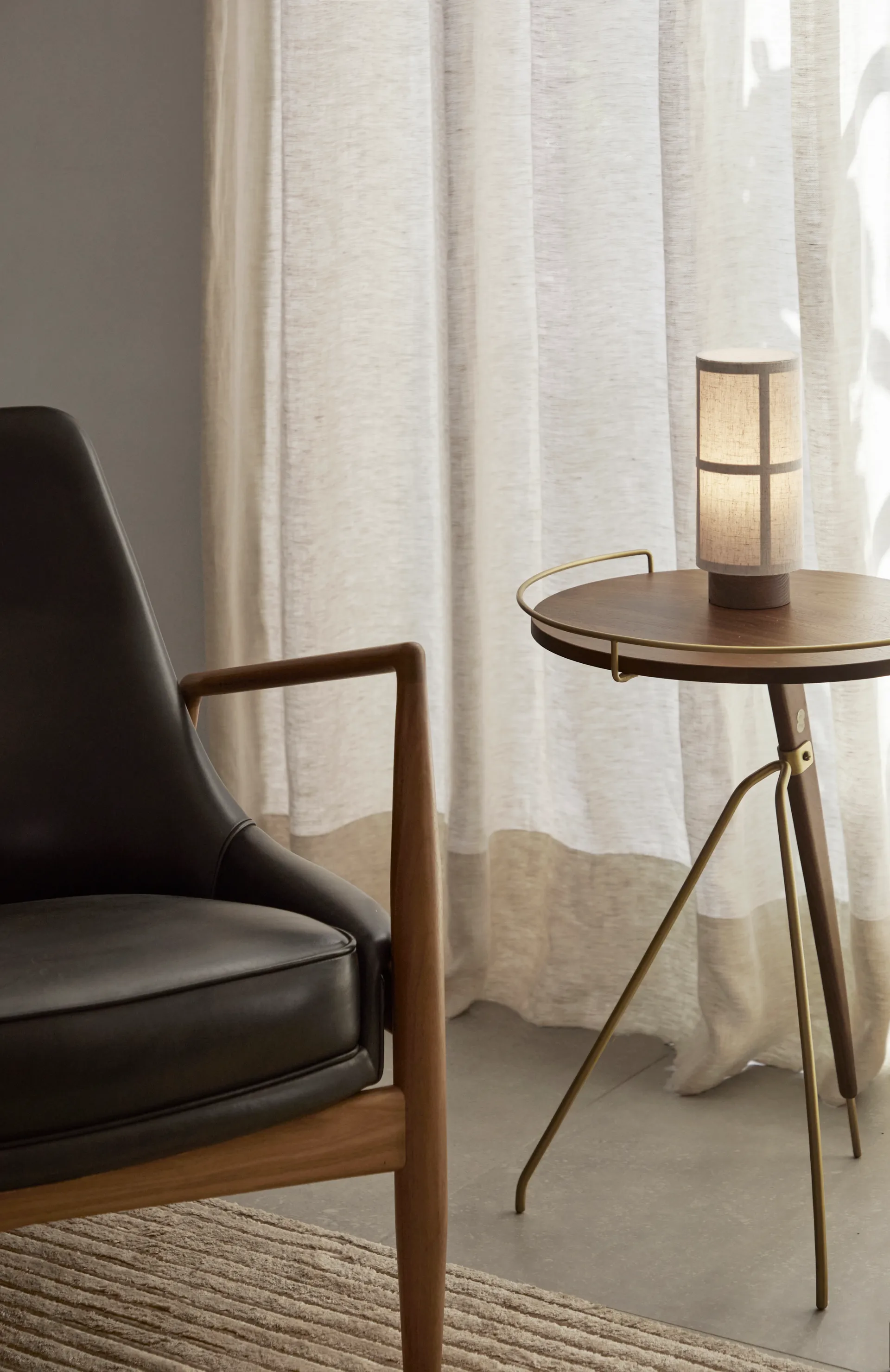 Audo Copenhagen black leather armchair, round wooden side table with a lit cylindrical lamp, and sheer curtains.