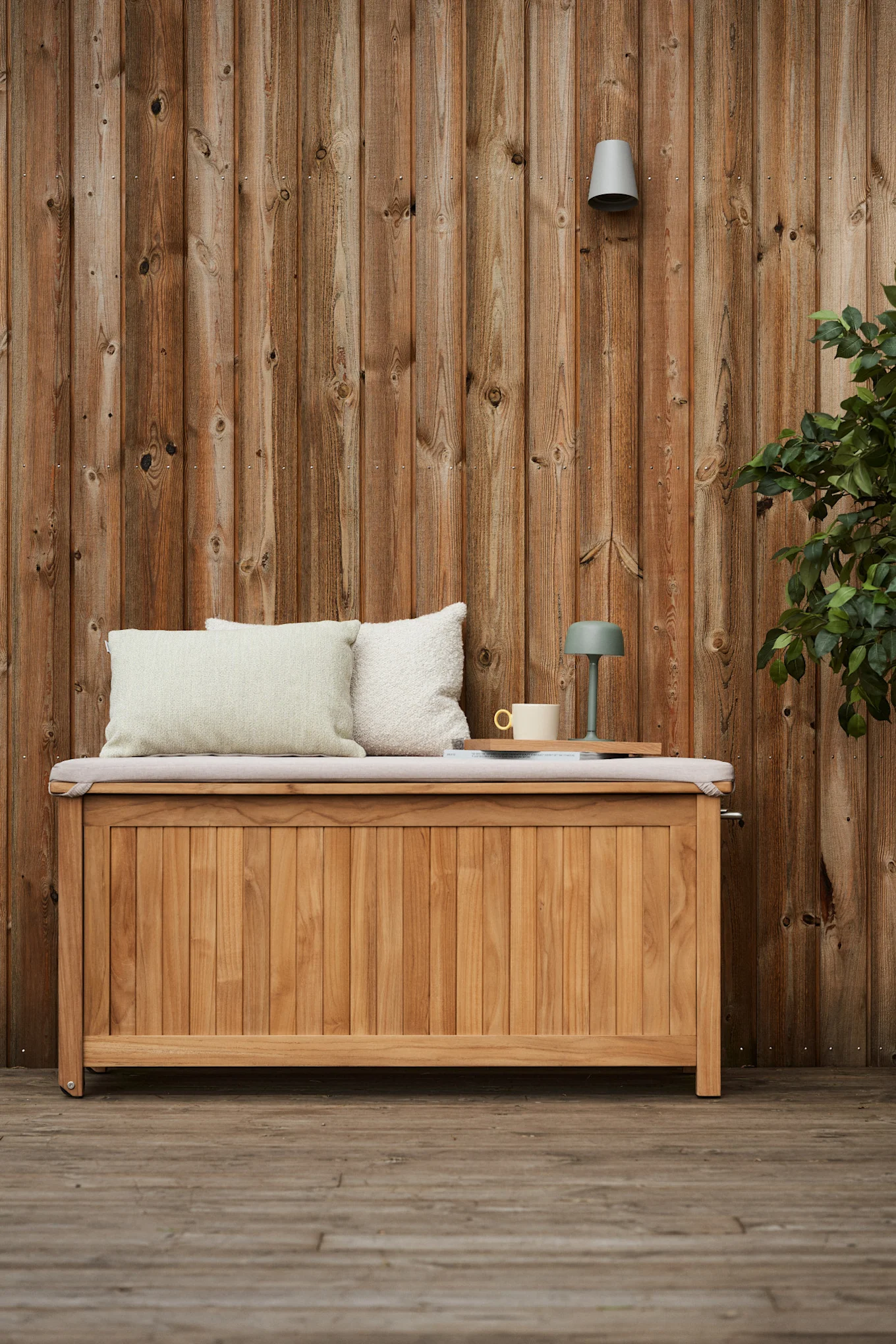 The Bjärknäs cushion box from 1898 stands on a terrace in front of a wooden house, with cosy cushions on top, a coffee cup and a portable table lamp next to it.