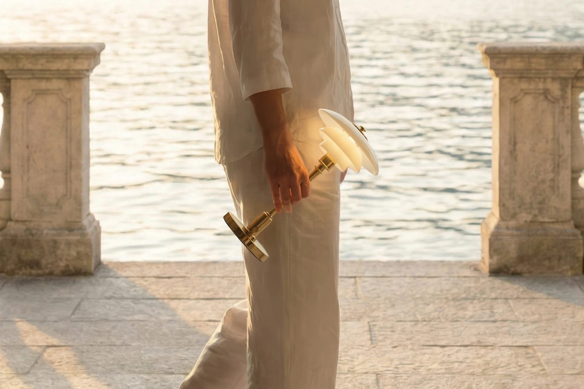 Person in white clothes holding an illuminated lamp with a brass base and layered white shades, walking by shimmering water with stone pillars.