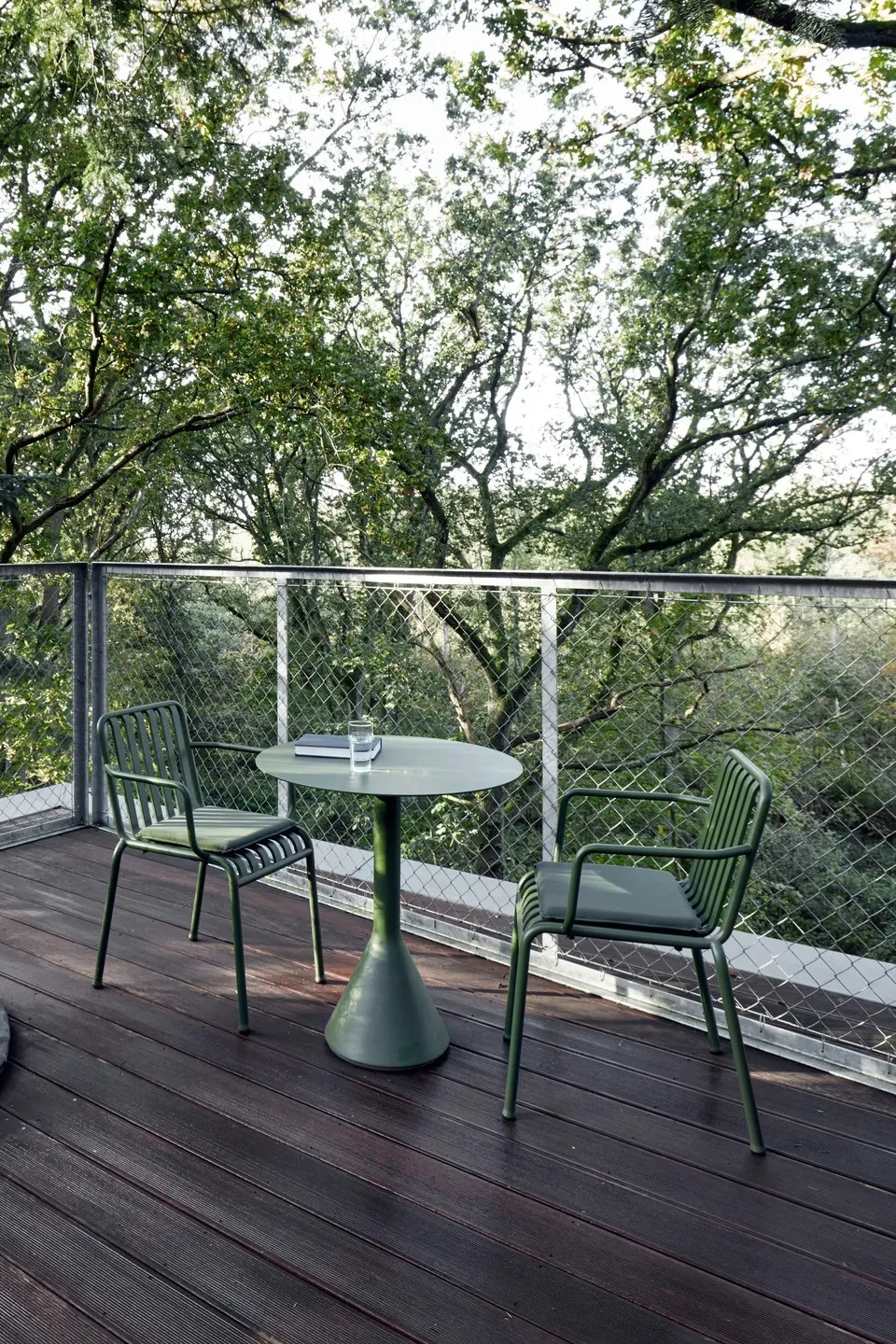 Two green outdoor chairs and a round table on a wooden deck, surrounded by lush trees.