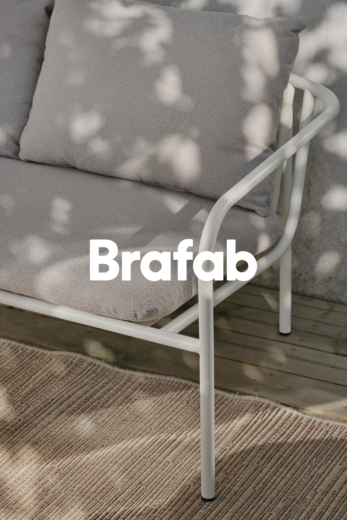 Brafab logo over a light grey outdoor sofa with a white metal frame, on a beige rug on wooden decking with dappled shadows.