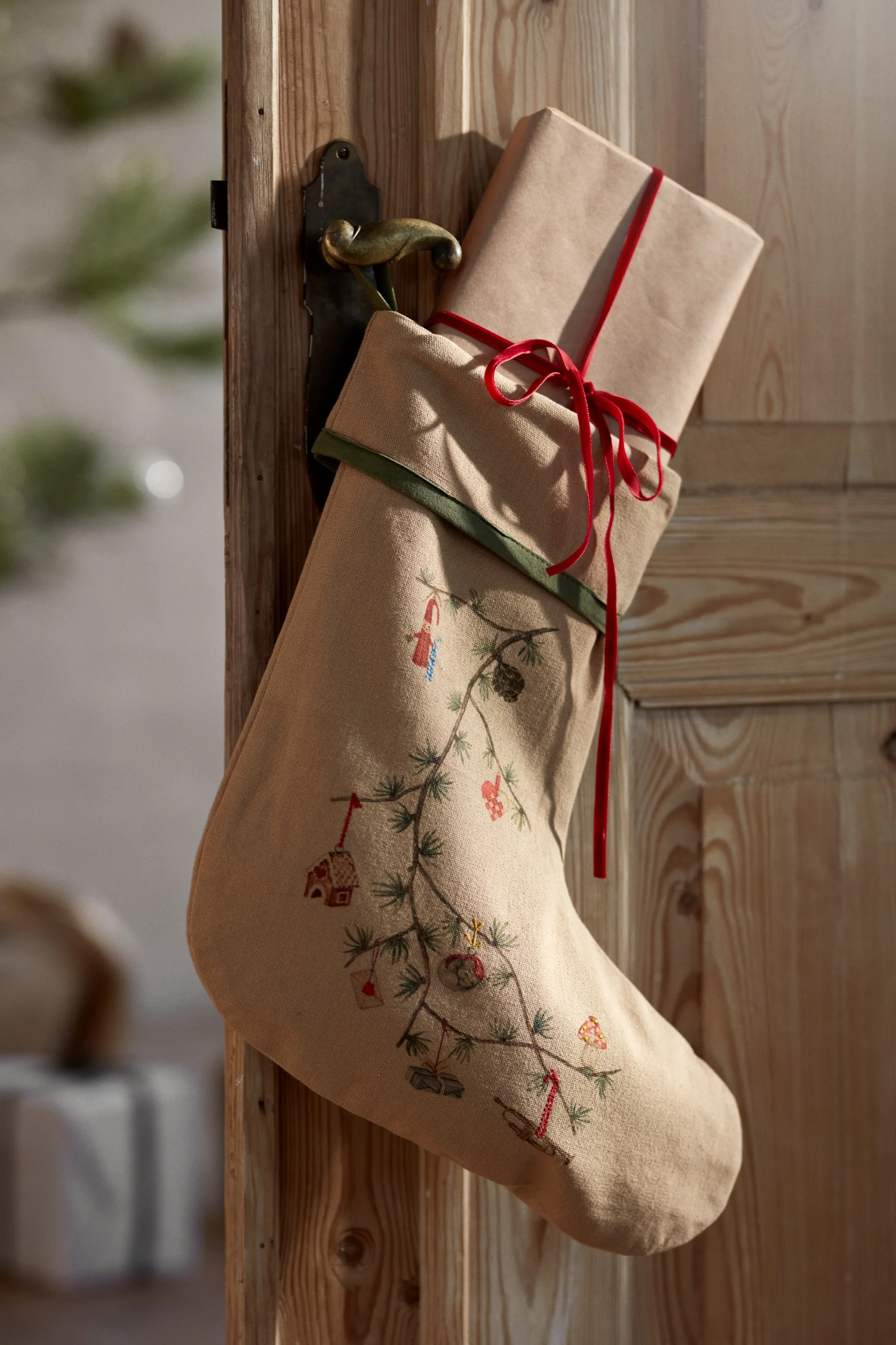 Christmas decorations for 2025 are inspired by nature like this stocking from Kähler that you see here hanging on a wooden door in front of a Christmas tree and wrapped gifts. 