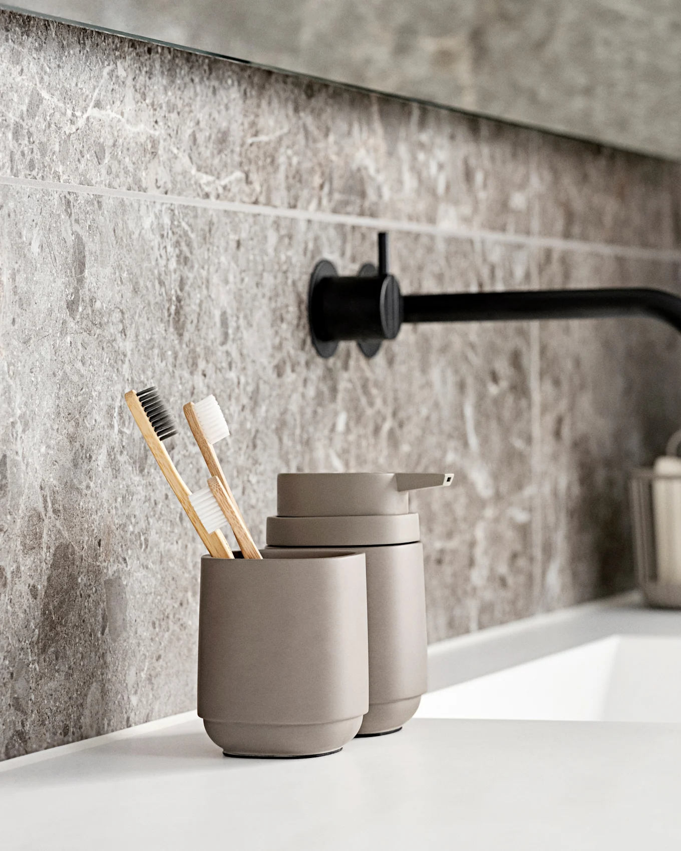 Modern bathroom with a white sink, black wall faucet, grey marble-look tiles, and eco-friendly toothbrushes.