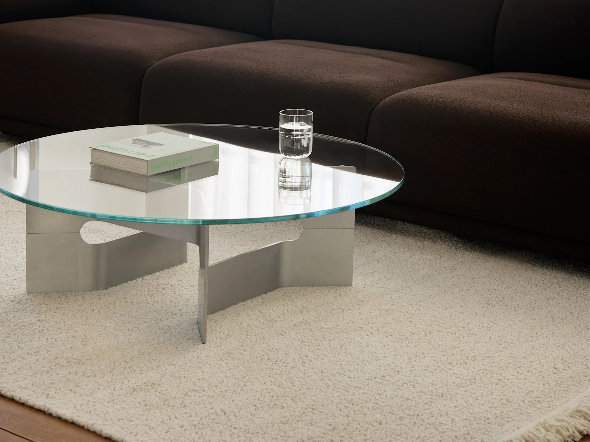 Round glass coffee table with books and a glass of water on a cream shaggy rug in front of a dark brown sofa.