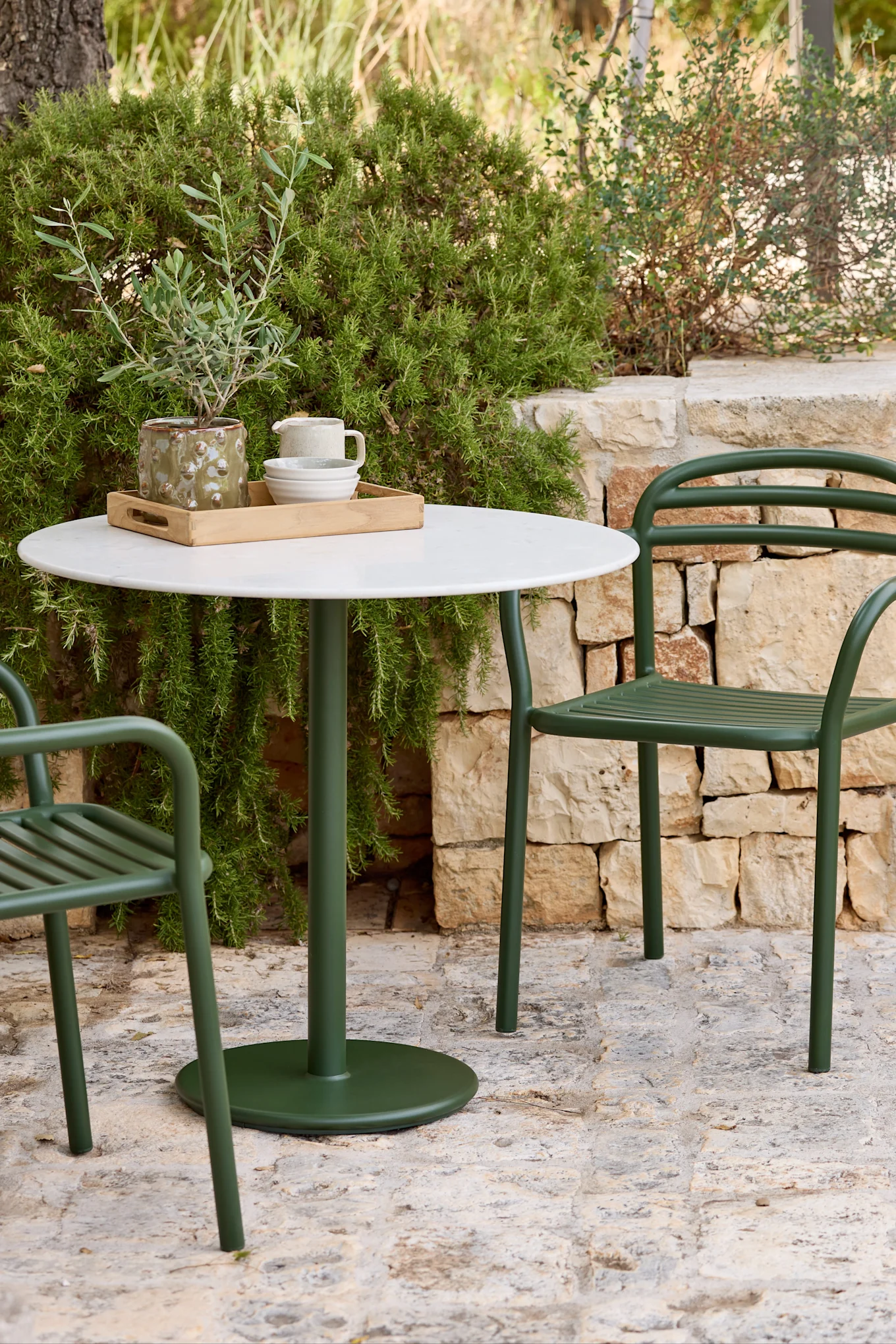 An outdoor dining set with a round white table and two dark green metal chairs on a stone patio, surrounded by lush green foliage.