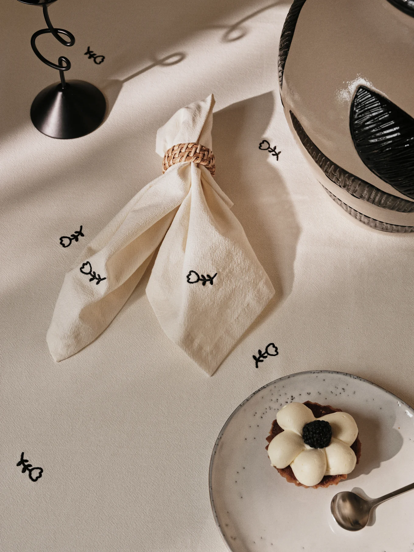 Table set with a cream napkin in a woven ring, black fish bone symbols, a coiled metal stand, and a blackberry tart on a speckled plate.