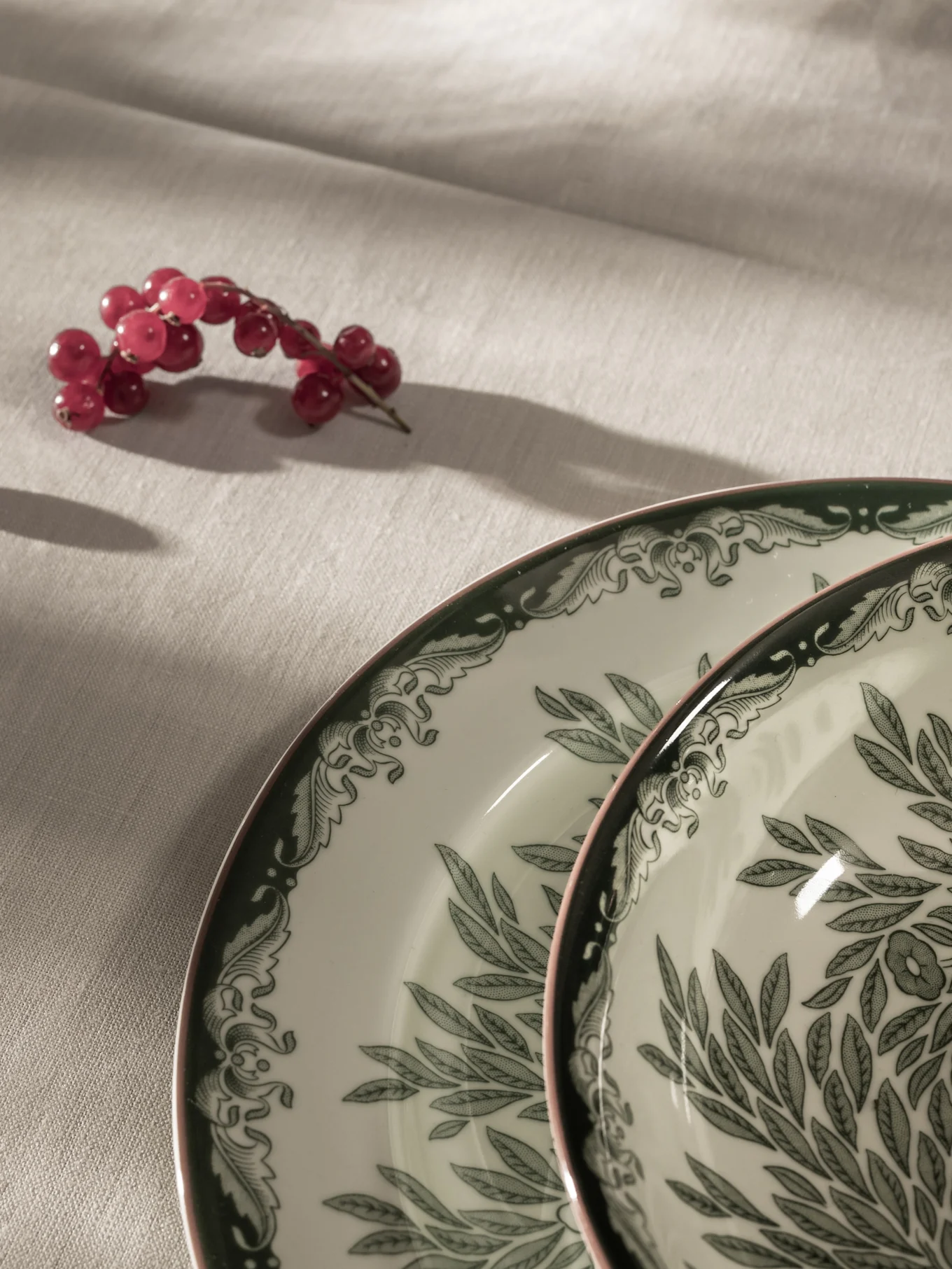Red currants on a light tablecloth next to two white plates with green leaf patterns and pink rims.