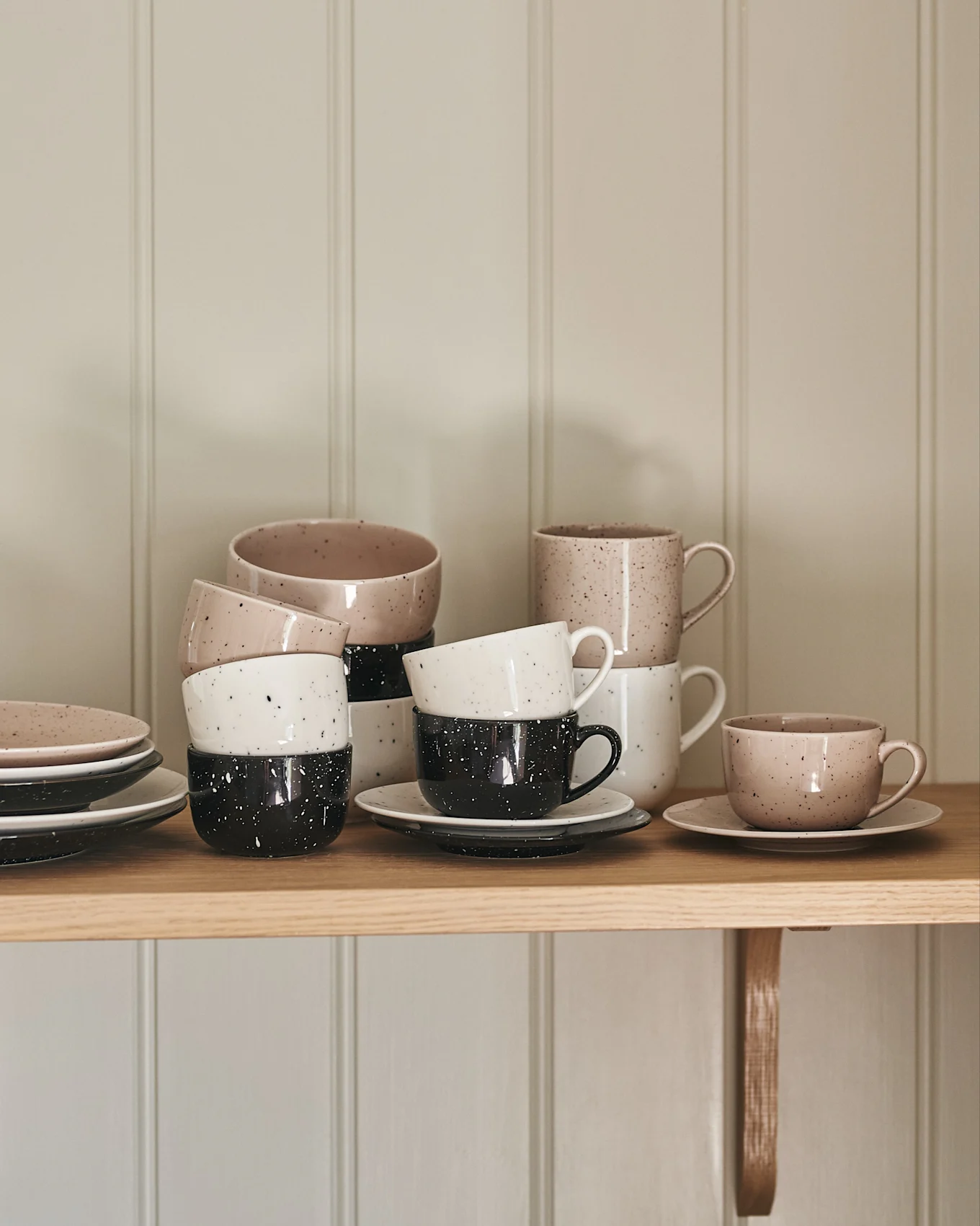 Stacked ceramic cups and plates in speckled beige, white, and black on a wooden shelf.