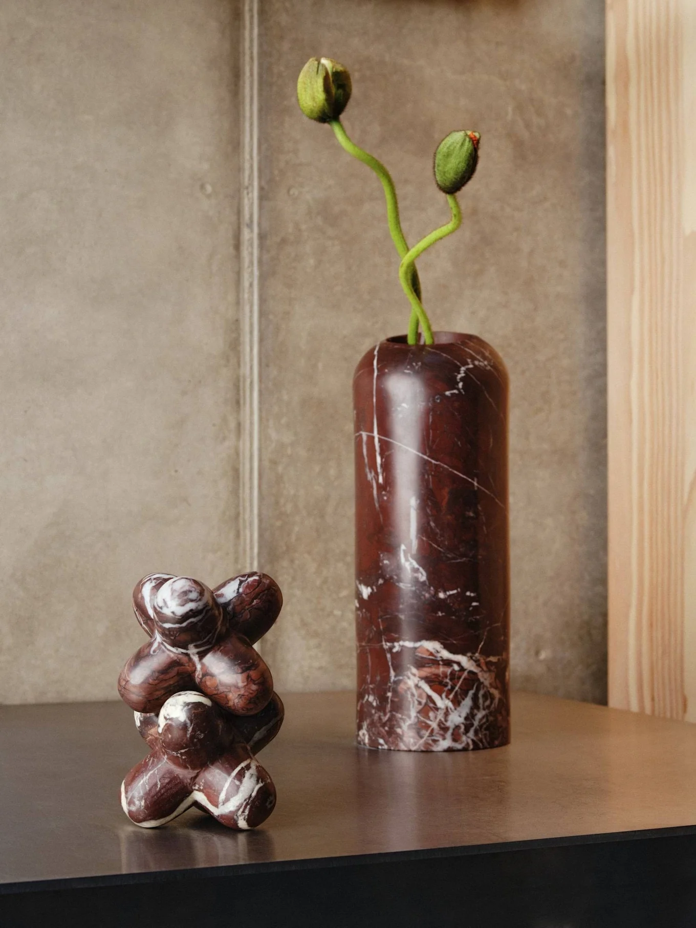 Two marble objects on a shelf: a tall dark red vase with green flower buds and an abstract sculptural piece.