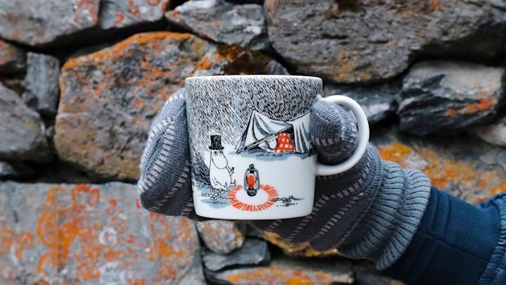 @moominbyingri holds the Schlaf gut Moomin cup in front of a stone wall wearing a glove.