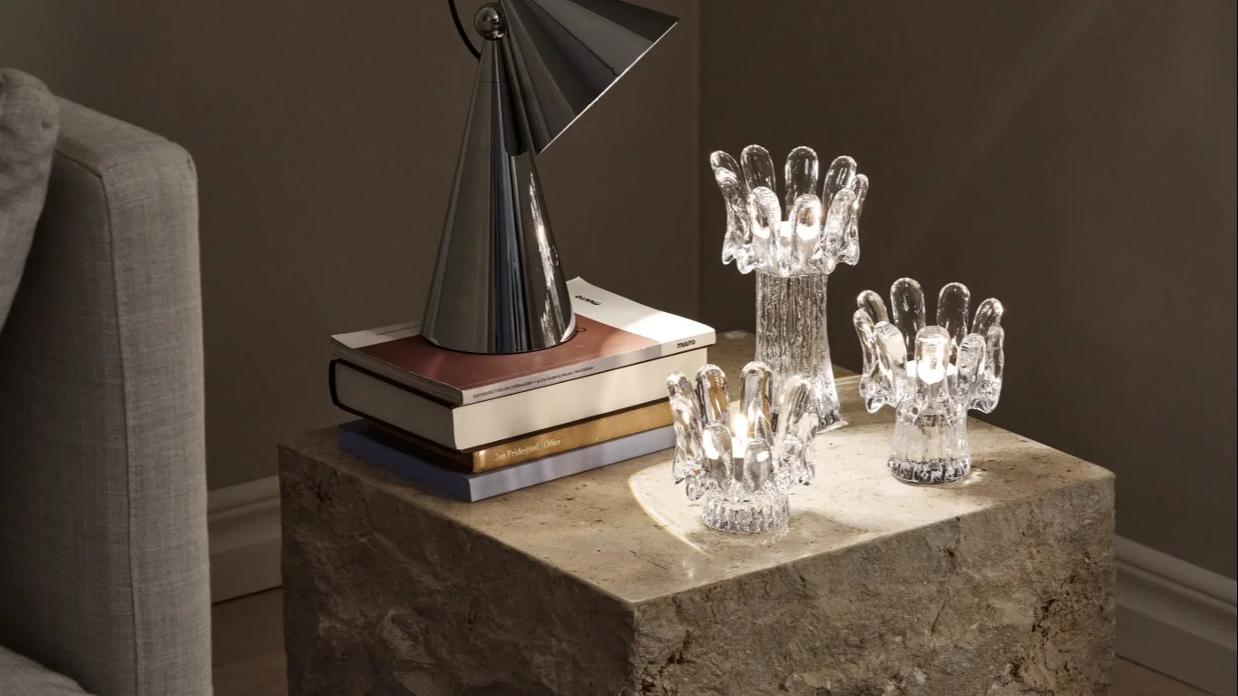 Three glass candle holders in different sizes sit on a stone table alongside a portable lamp and books, together reflecting the interior Scandinavian design trends for 2026.