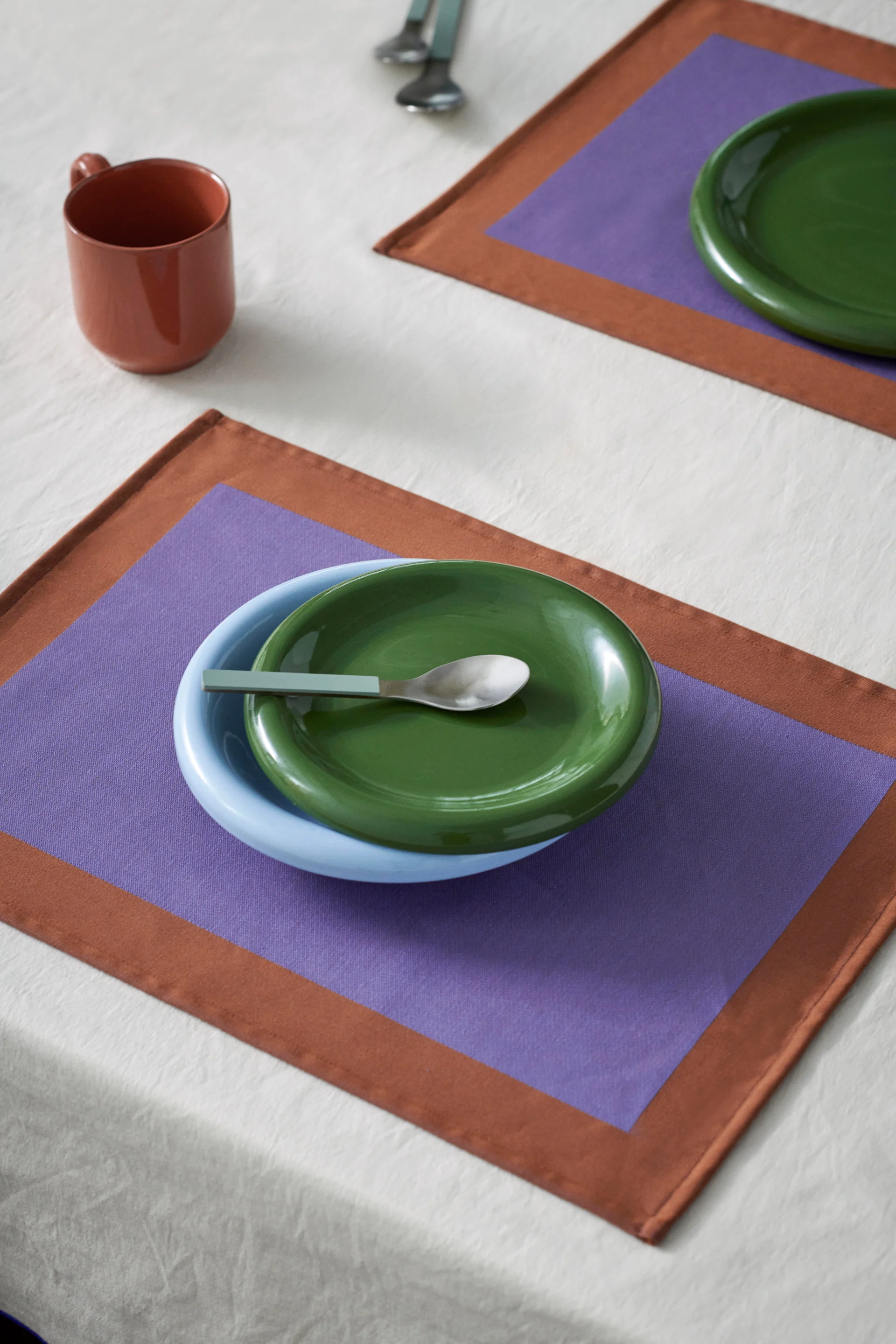 A table setting with a blue bowl, green plate, and a spoon on a purple and brown placemat. A brown mug is also on the white tablecloth.