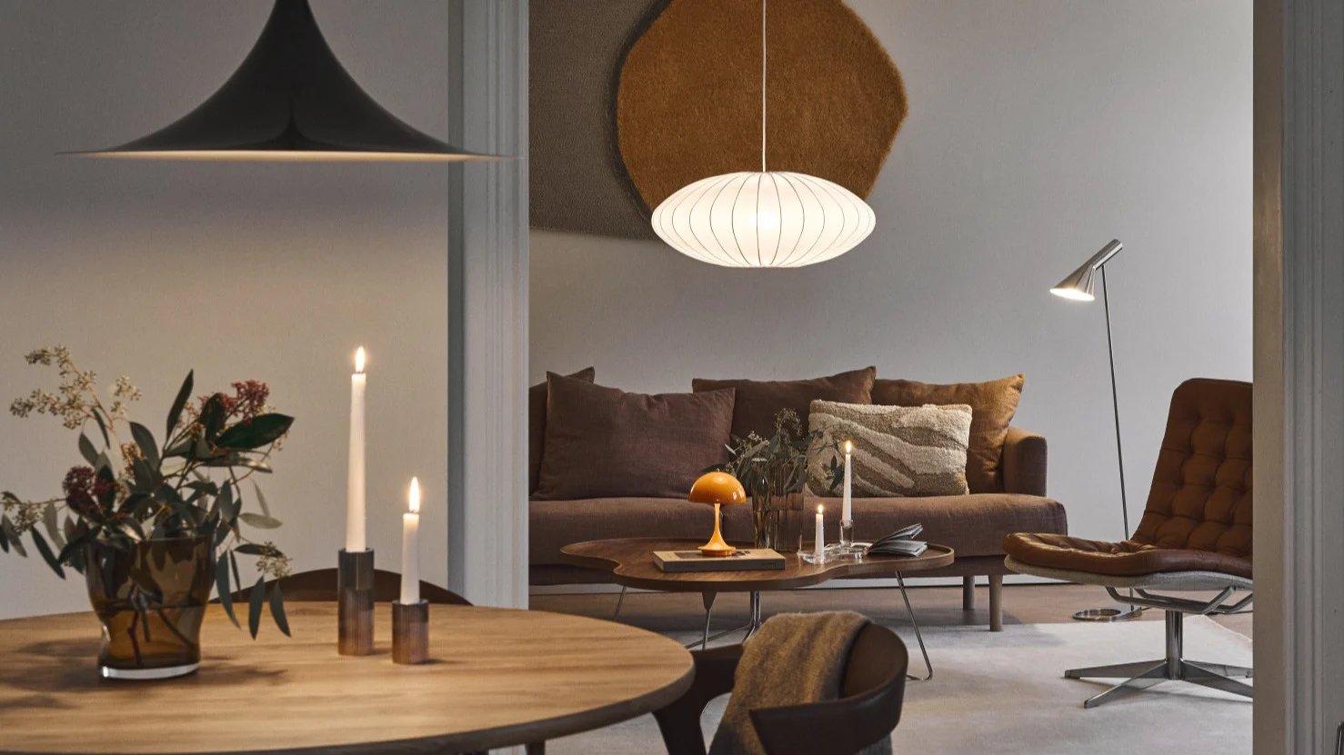 A warmly lit modern dining and living room featuring varied designer lamps, a brown sofa, and a tan leather lounge chair.