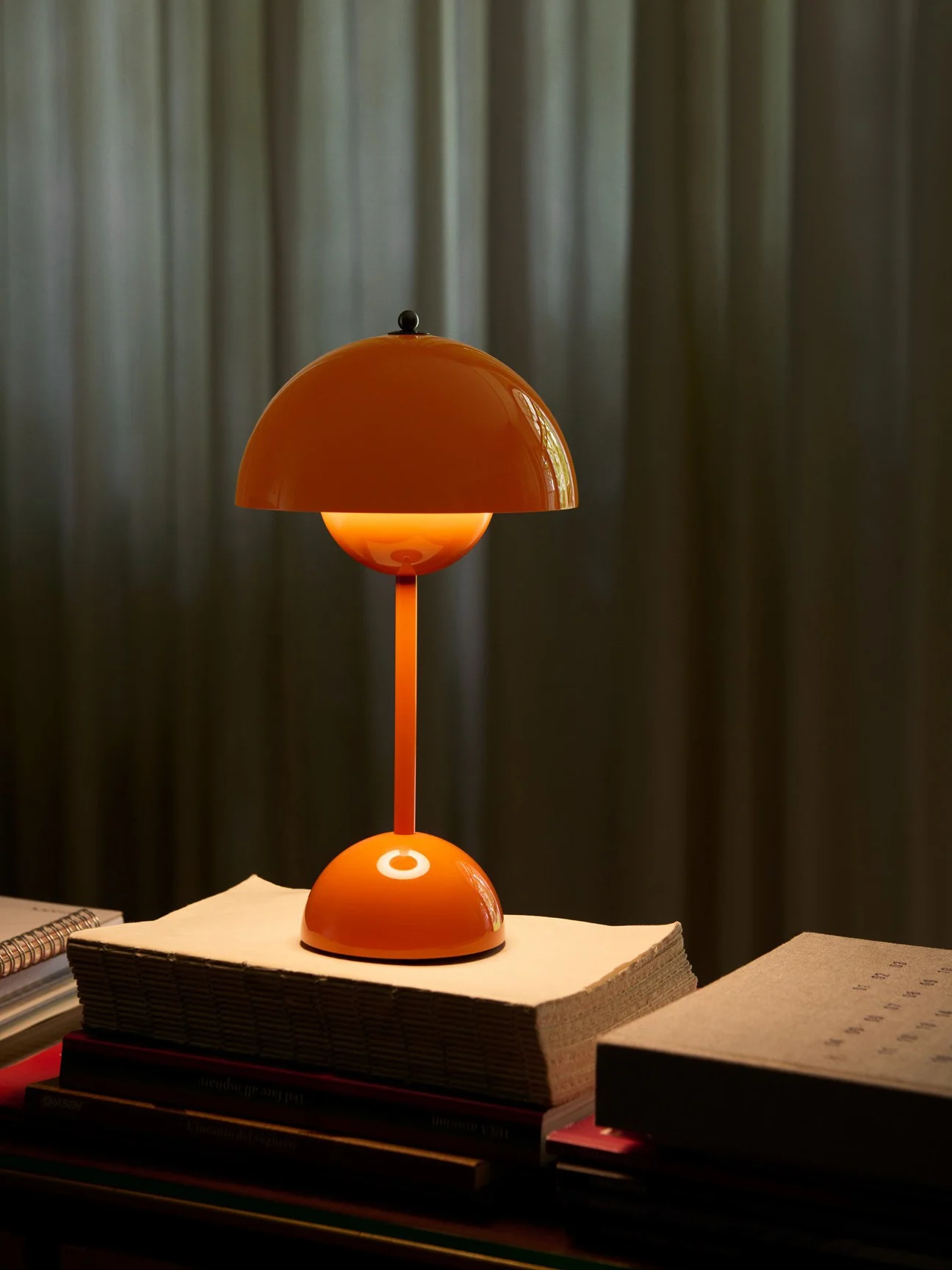 Bright orange dome-shaped table lamp on a stack of books, casting a warm glow against a dark curtain.