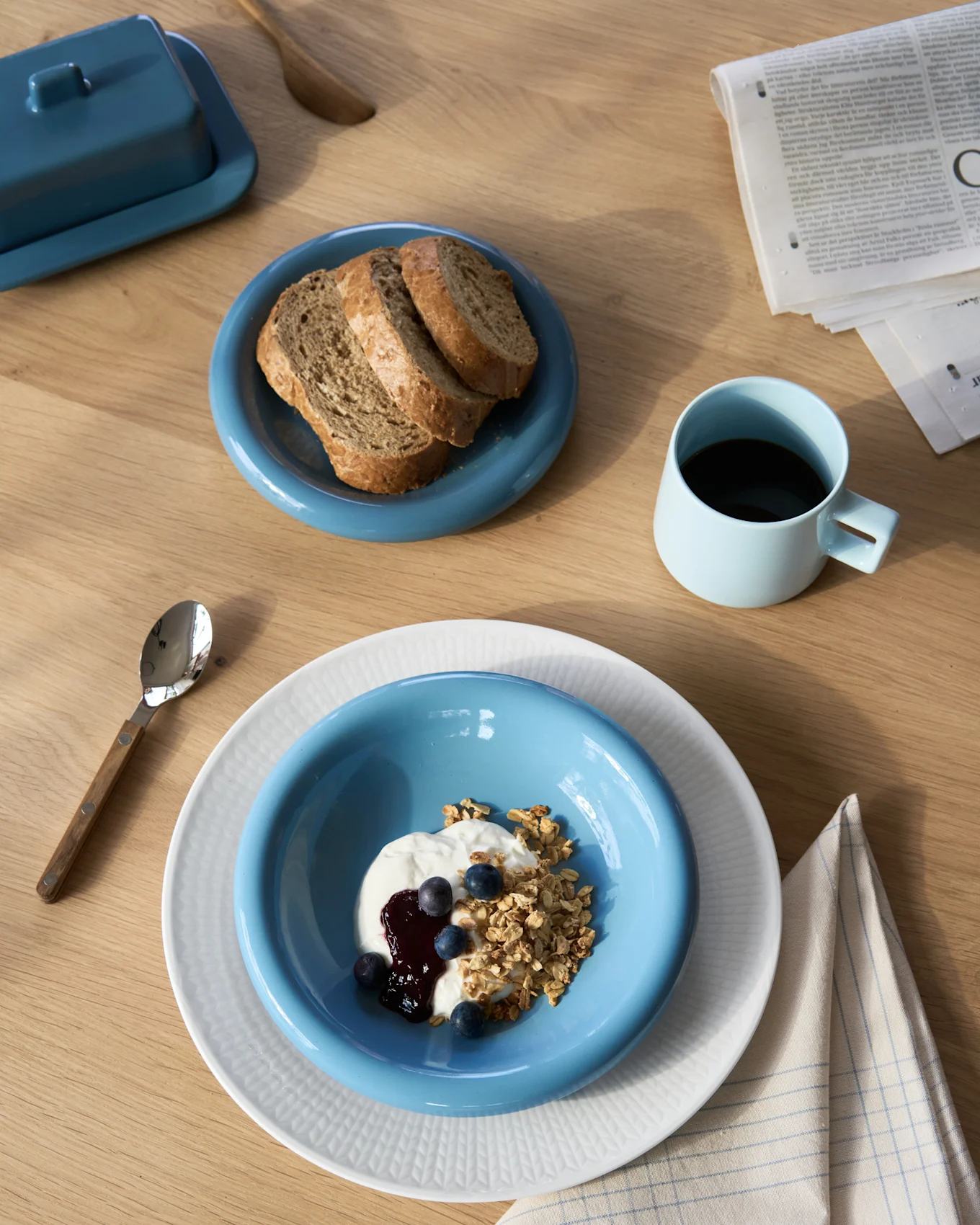 Overhead view of a breakfast spread with yogurt and granola, dark bread, coffee, and a butter dish on a wooden table.