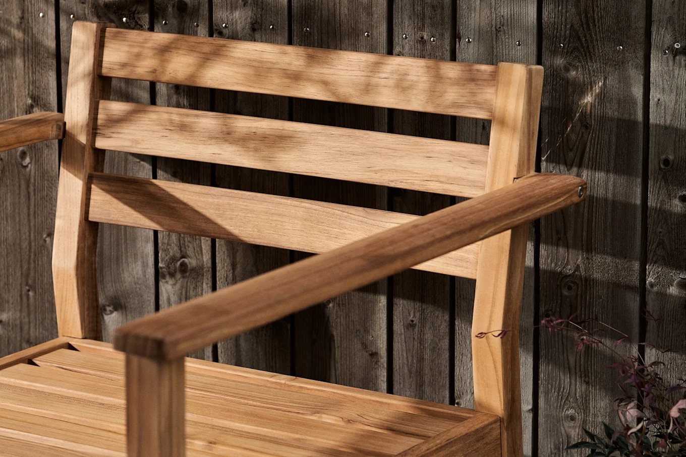 Slatted wooden chair with armrests against a dark wooden fence, covered in leaf shadows.