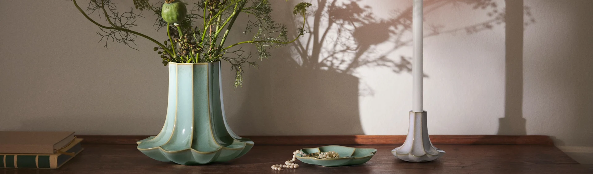 A fluted teal vase with foliage, a pearl necklace in a matching dish, a white candle in a fluted holder, and books on a wooden surface.