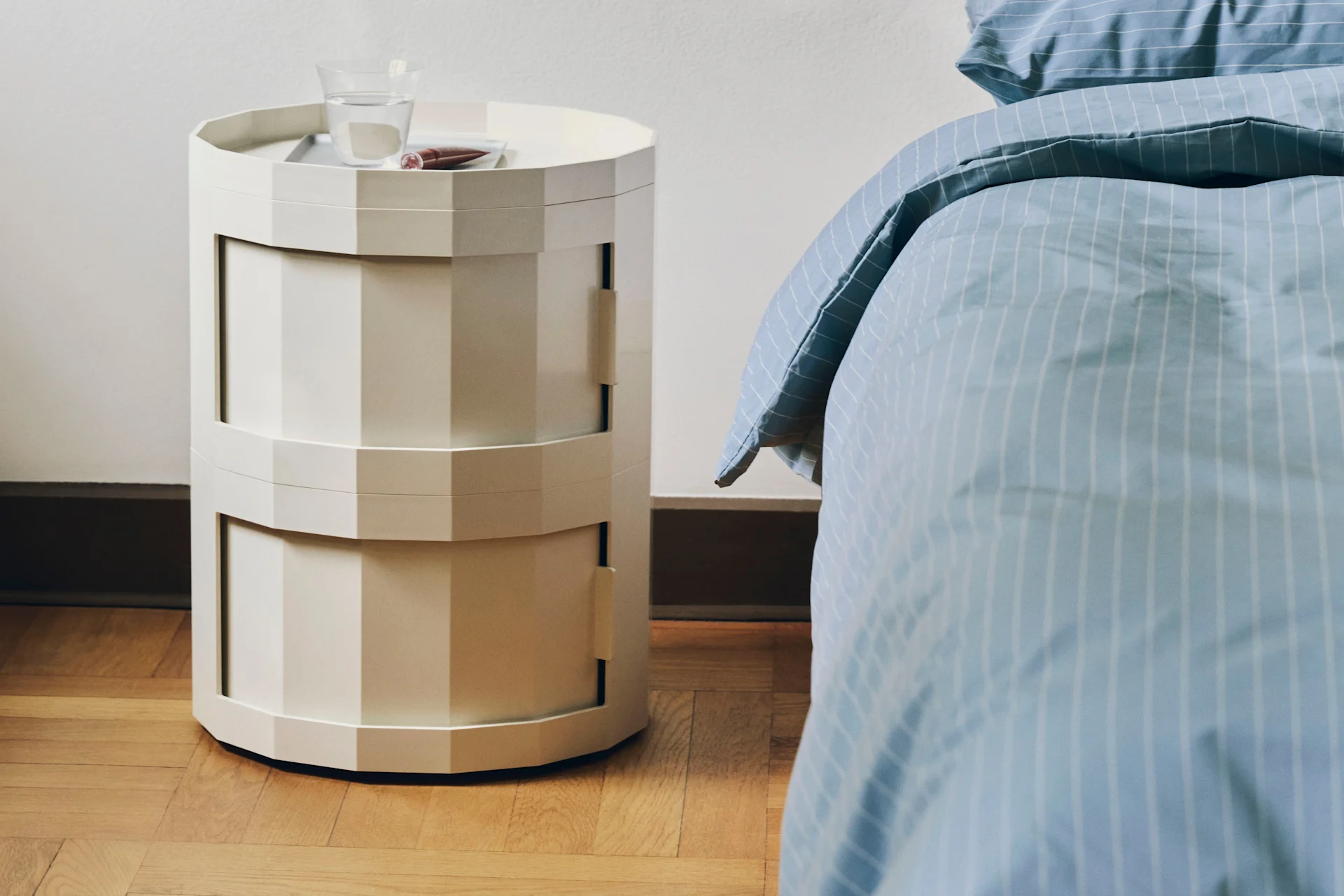 A light-colored, hexagonal, multi-tiered nightstand with a glass of water and cosmetic on top, next to a bed with blue striped bedding.