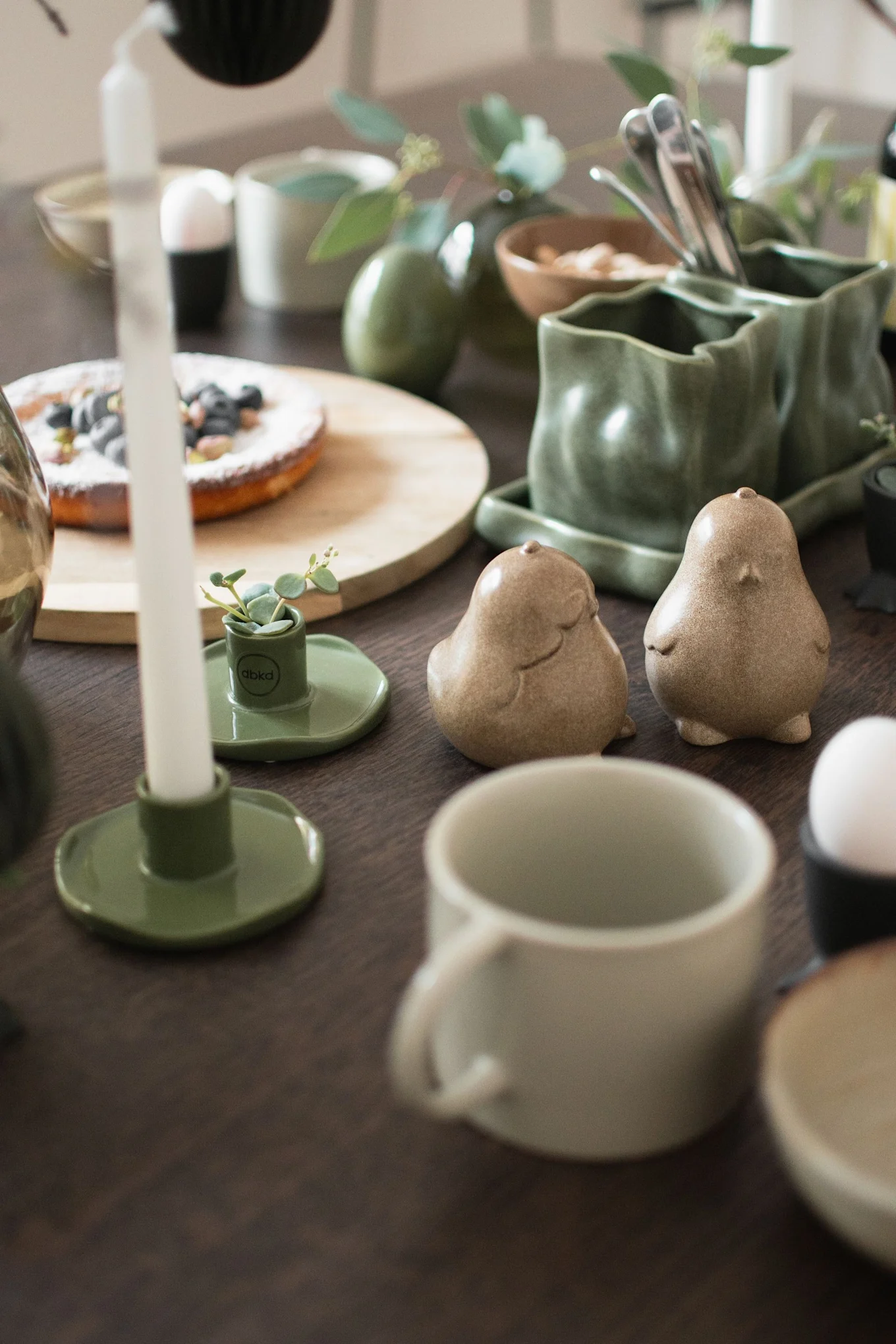 Table set with green ceramic dishware, a blueberry tart, candles, and two bird-shaped shakers.