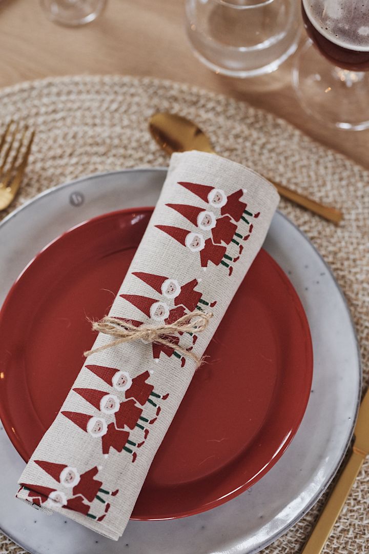 A rolled up tea towel with Santa Claus motifs rests on a red plate and a Nordic sand plate from Broste Copenhagen.