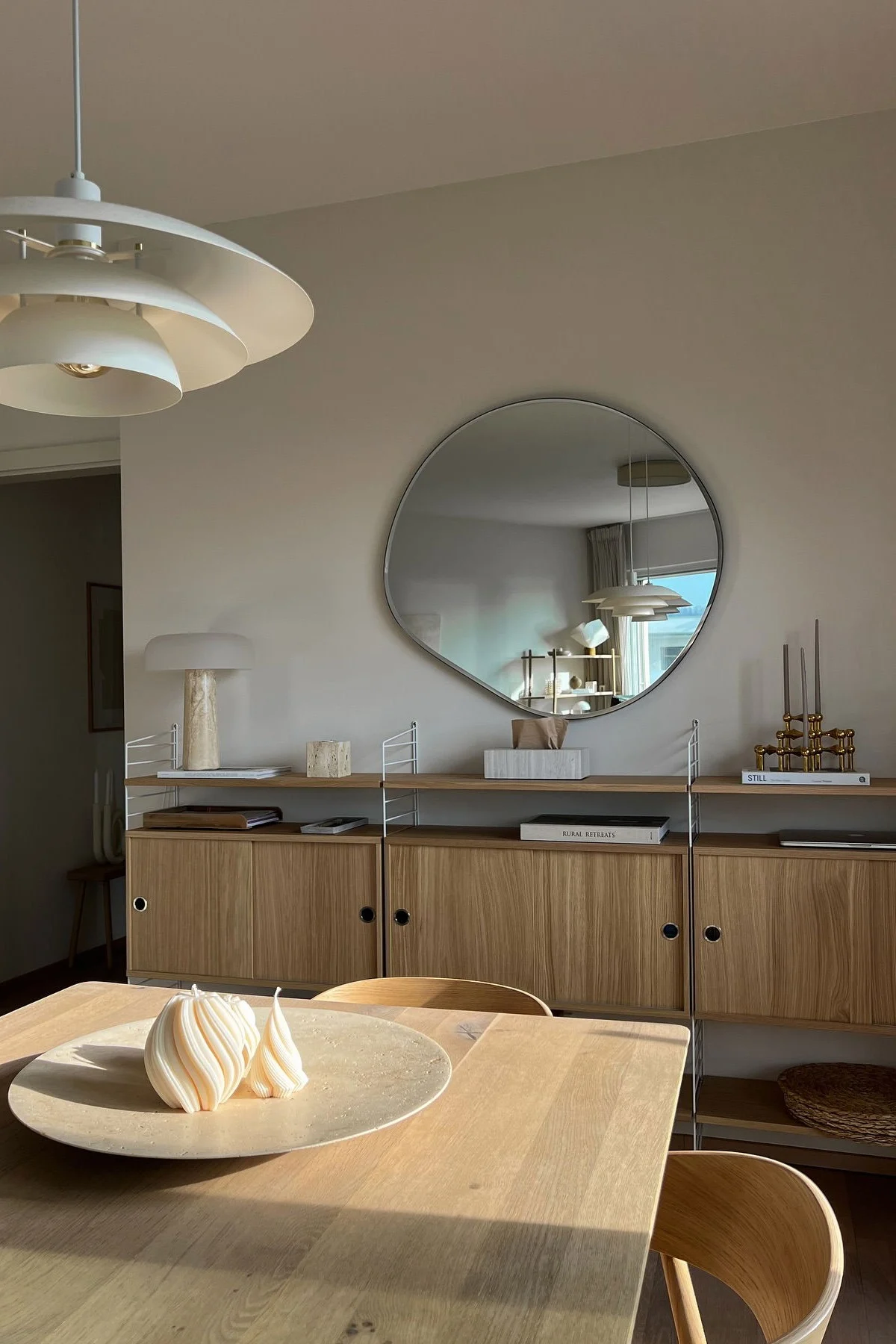 Modern dining room with light wood table, two white spiral candles, wood shelf, irregular wall mirror, and tiered white lamp.