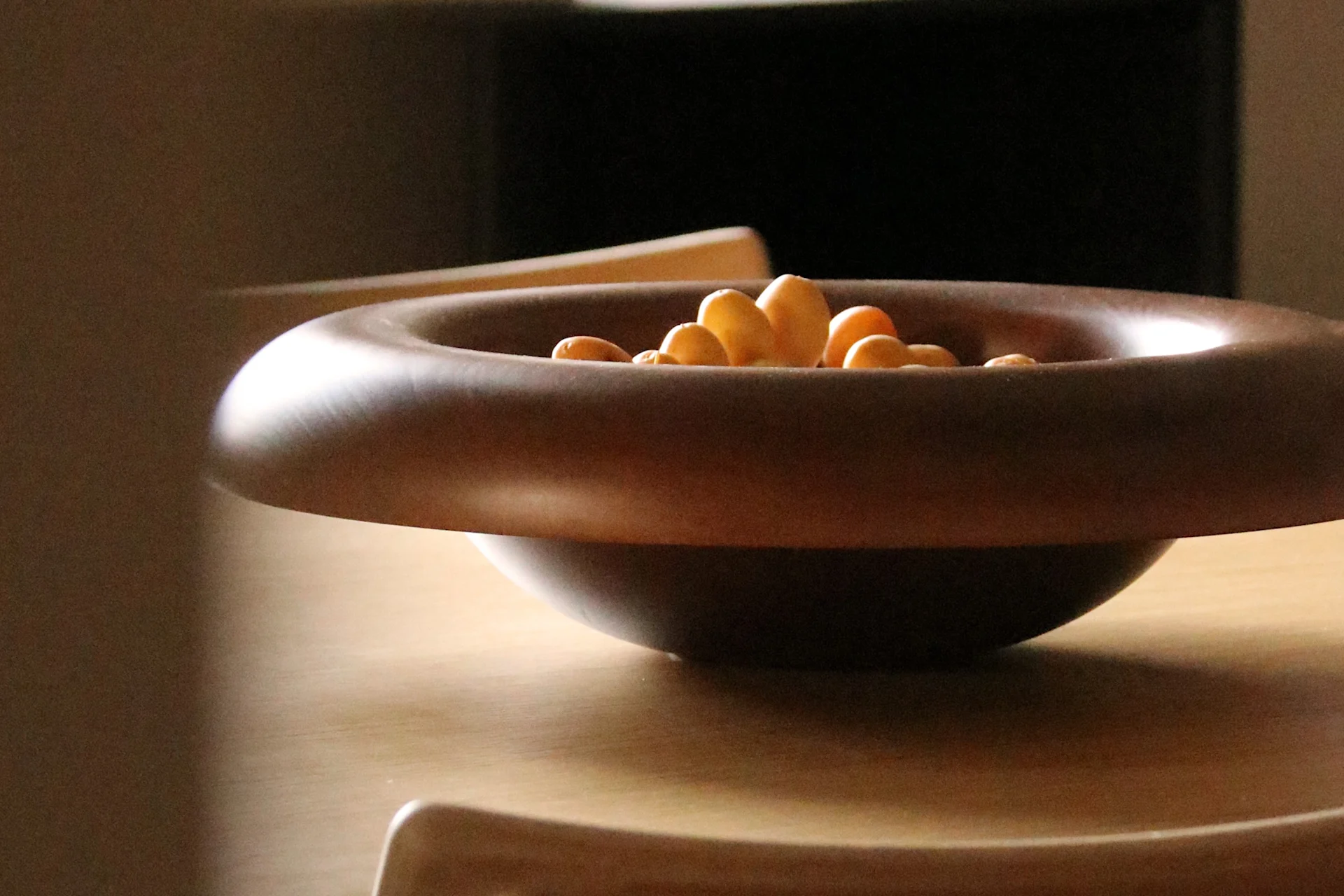 Dark brown wooden bowl with a wide rim filled with light-colored snacks on a light wooden table.