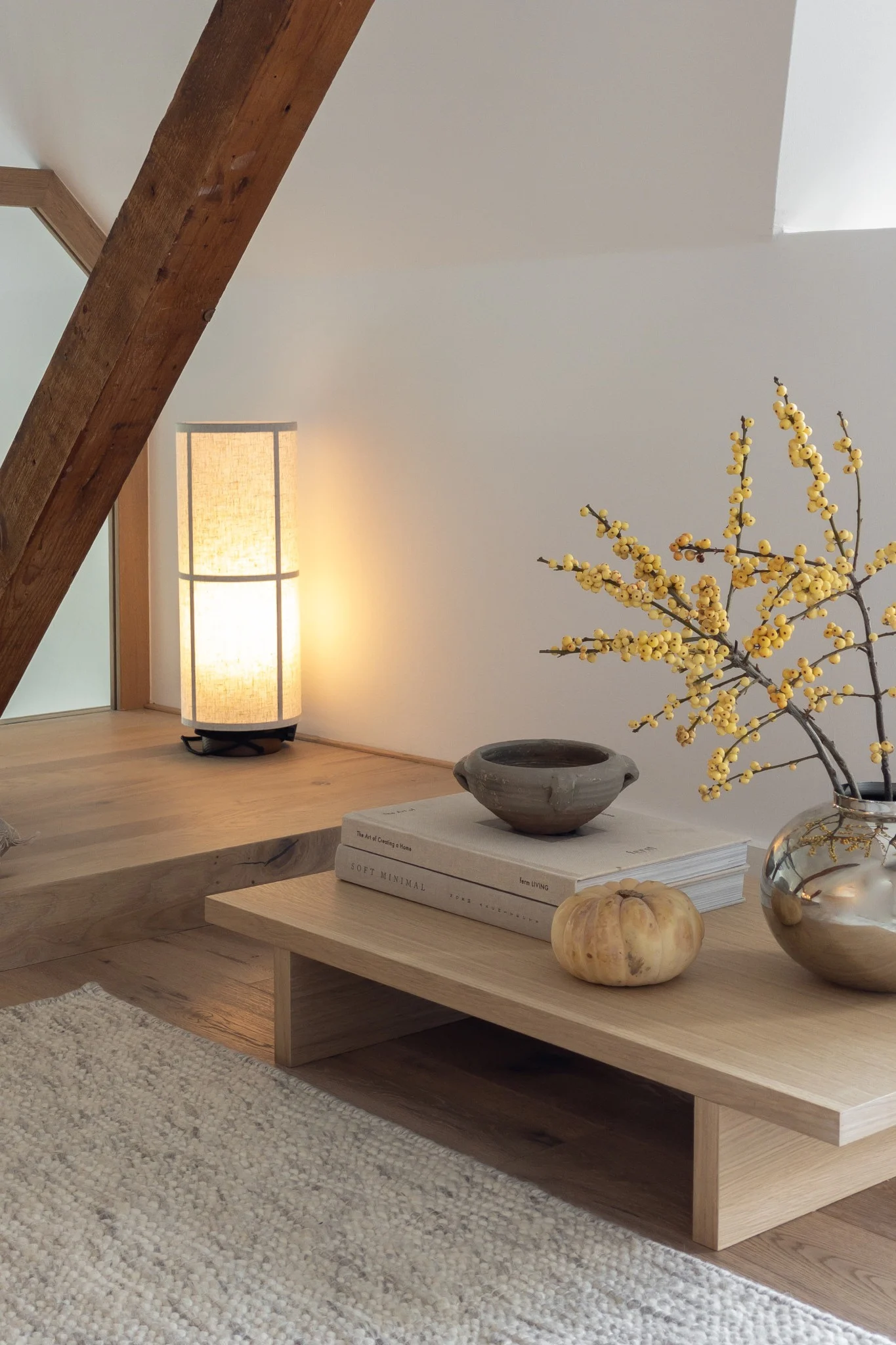 A minimalist room with a tall, rectangular lamp glowing warmly next to a low wood coffee table adorned with books, a rustic bowl, a small pumpkin, and a vase of yellow berries.