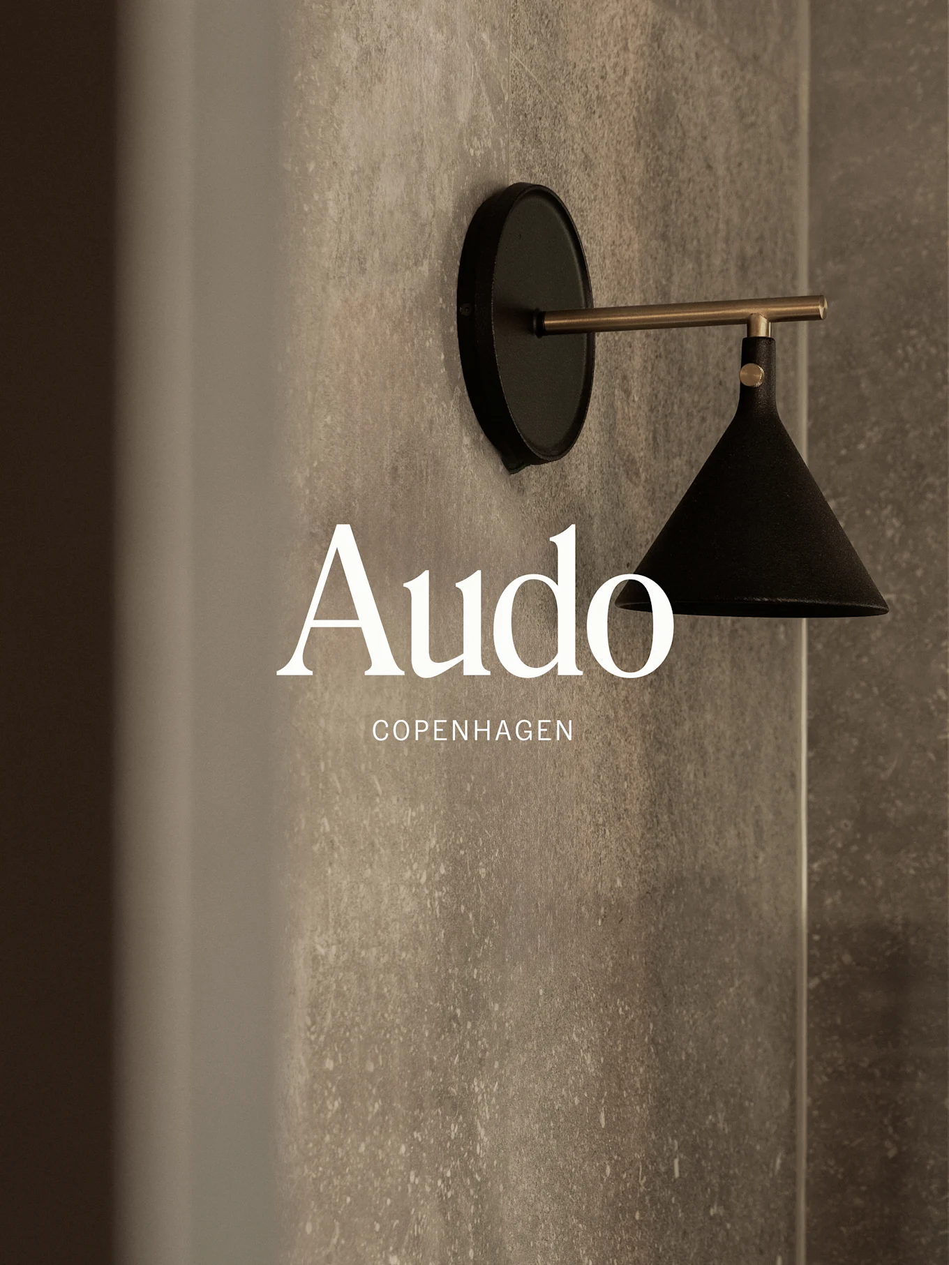 A wall-mounted black lamp with a brass arm against a textured grey wall, with 'Audo Copenhagen' text.