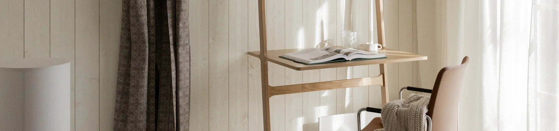 A wooden stand with an open book, glass vase, and white mug, next to a brown leather chair with a beige blanket, bathed in sunlight.