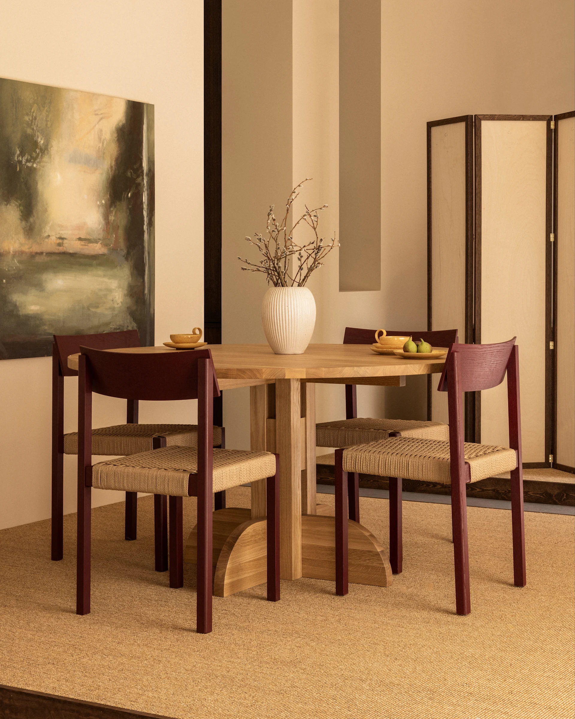 A dining room with a round wooden table, four chairs with reddish-brown frames and woven seats, and a vase of pussy willows.