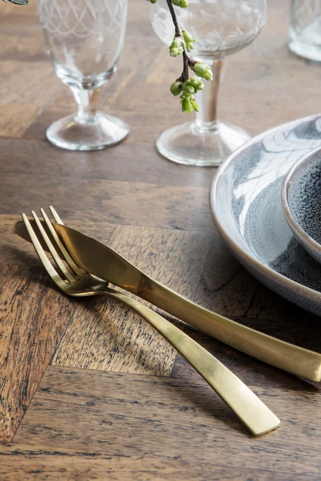 Table setting with stacked grey ceramic plates, gold cutlery, clear etched glasses, and a branch with green buds.