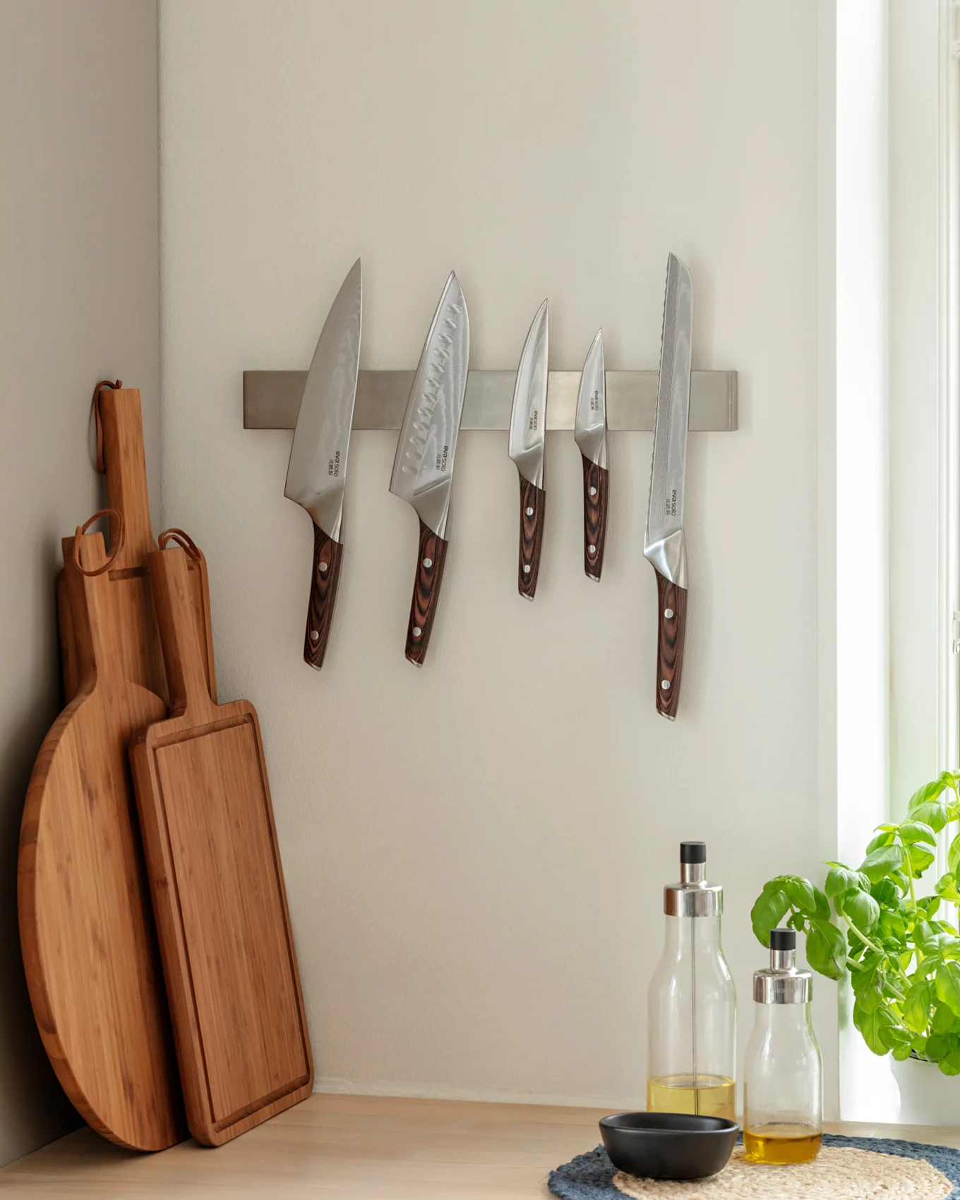 Kitchen corner with five knives on a magnetic strip, two wooden cutting boards, and two bottles of olive oil on a wooden counter next to a basil plant.
