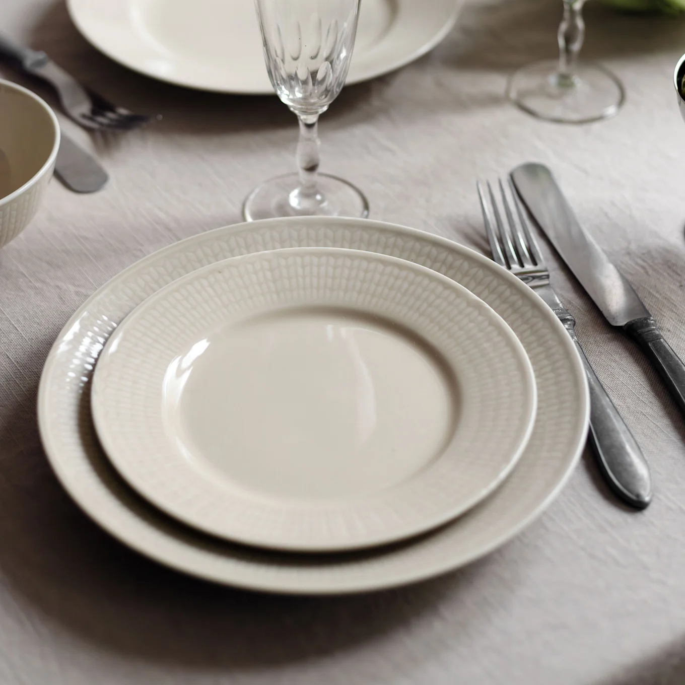 Two stacked cream plates with a textured rim, a fork, and a knife on a linen tablecloth.