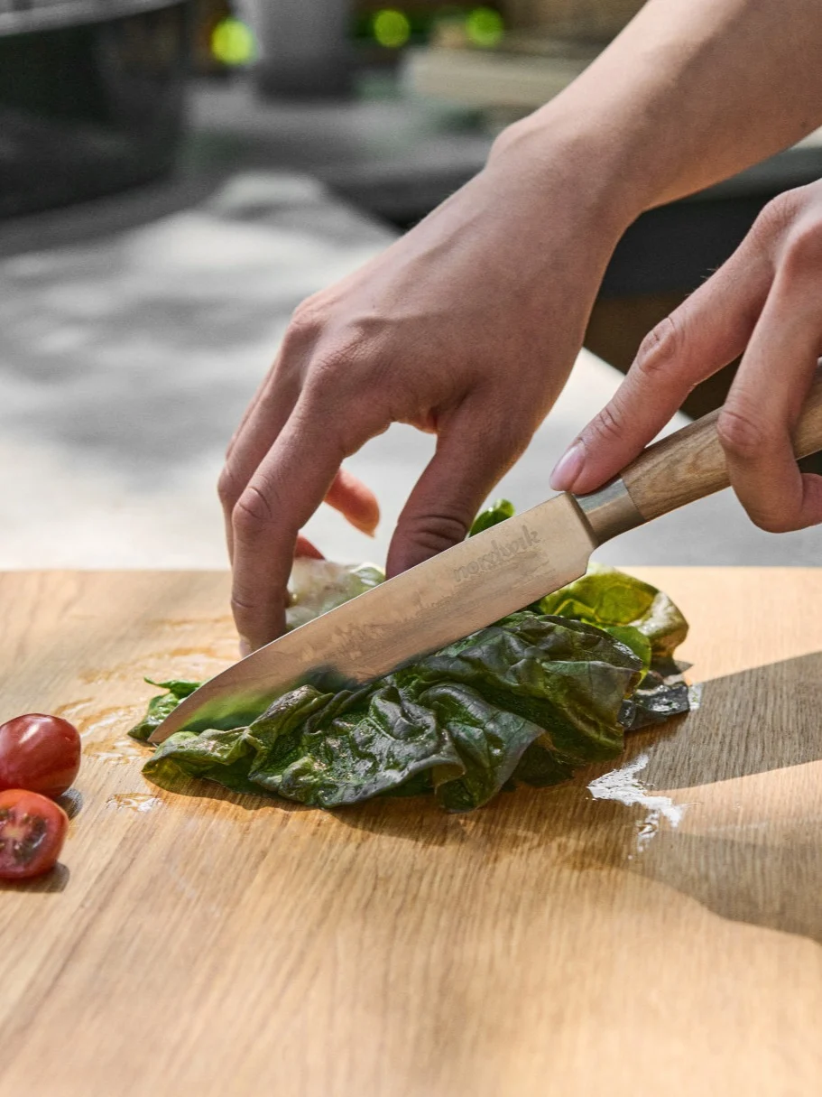 Hands chopping fresh green lettuce on a wooden board with a knife, alongside sliced cherry tomatoes.