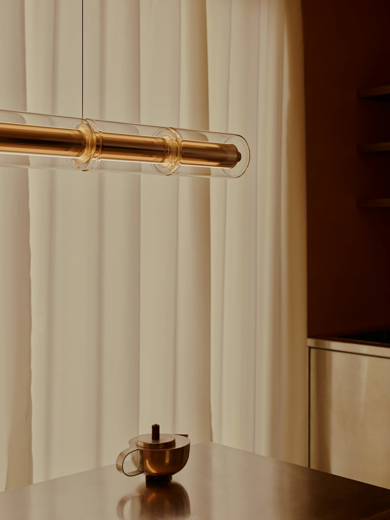 A contemporary brass and glass tube pendant light hangs above a brass teapot on a metallic table, with light curtains in the background.
