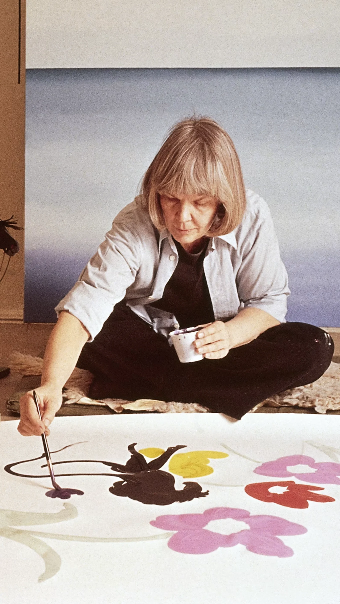 Artist sitting cross-legged, painting abstract colorful floral shapes on a large canvas on the floor.