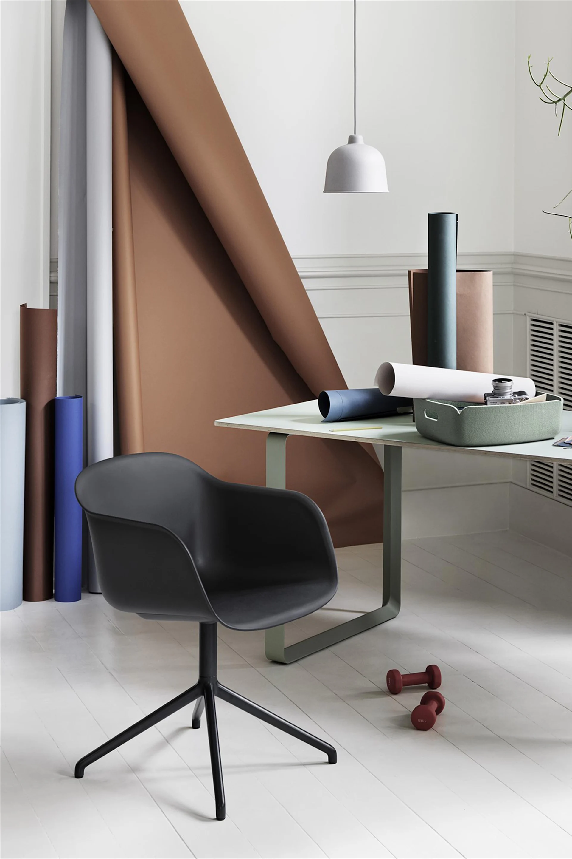 A black office chair in a bright room with a light green desk, rolls of colored paper, and two red dumbbells on the white floor.