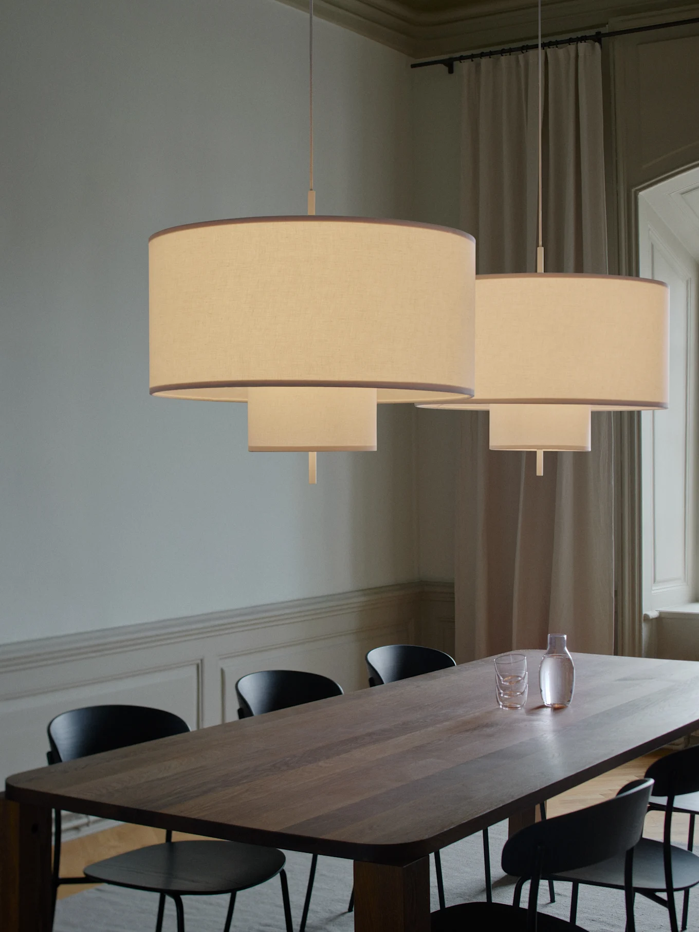Two tiered fabric pendant lights illuminate a long wooden dining table with black chairs, a carafe, and glasses.