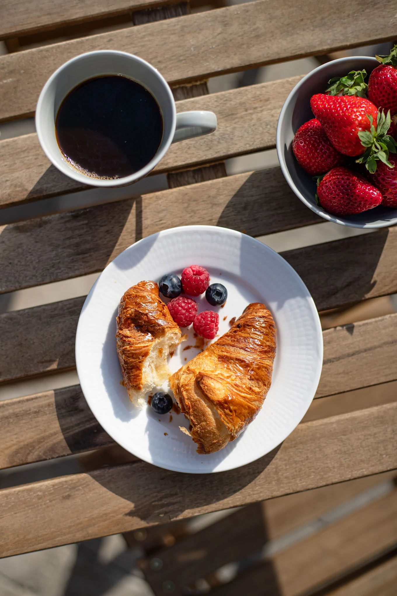 Overhead view of a breakfast spread on a wooden table with coffee, strawberries, croissants, blueberries, and raspberries.
