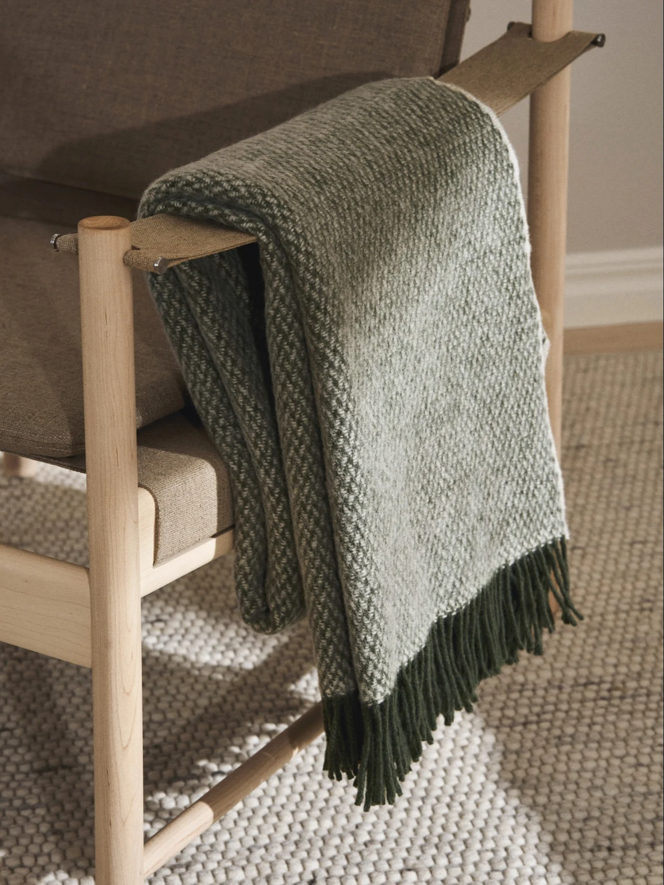 A light brown wooden armchair with a green and white patterned throw blanket draped over its armrest, featuring dark green fringes.