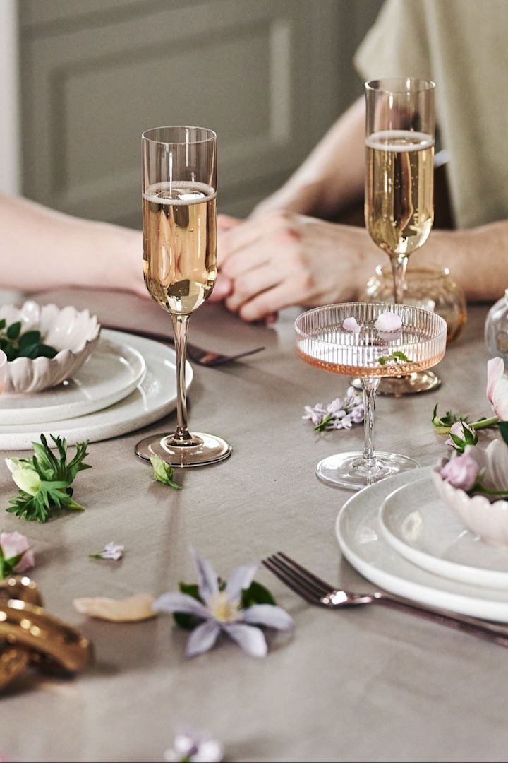 You can also decorate the table in a playful way for your romantic meal at home. Here you can see white crockery, simple glasses and flowers on a beige-coloured tablecloth.
